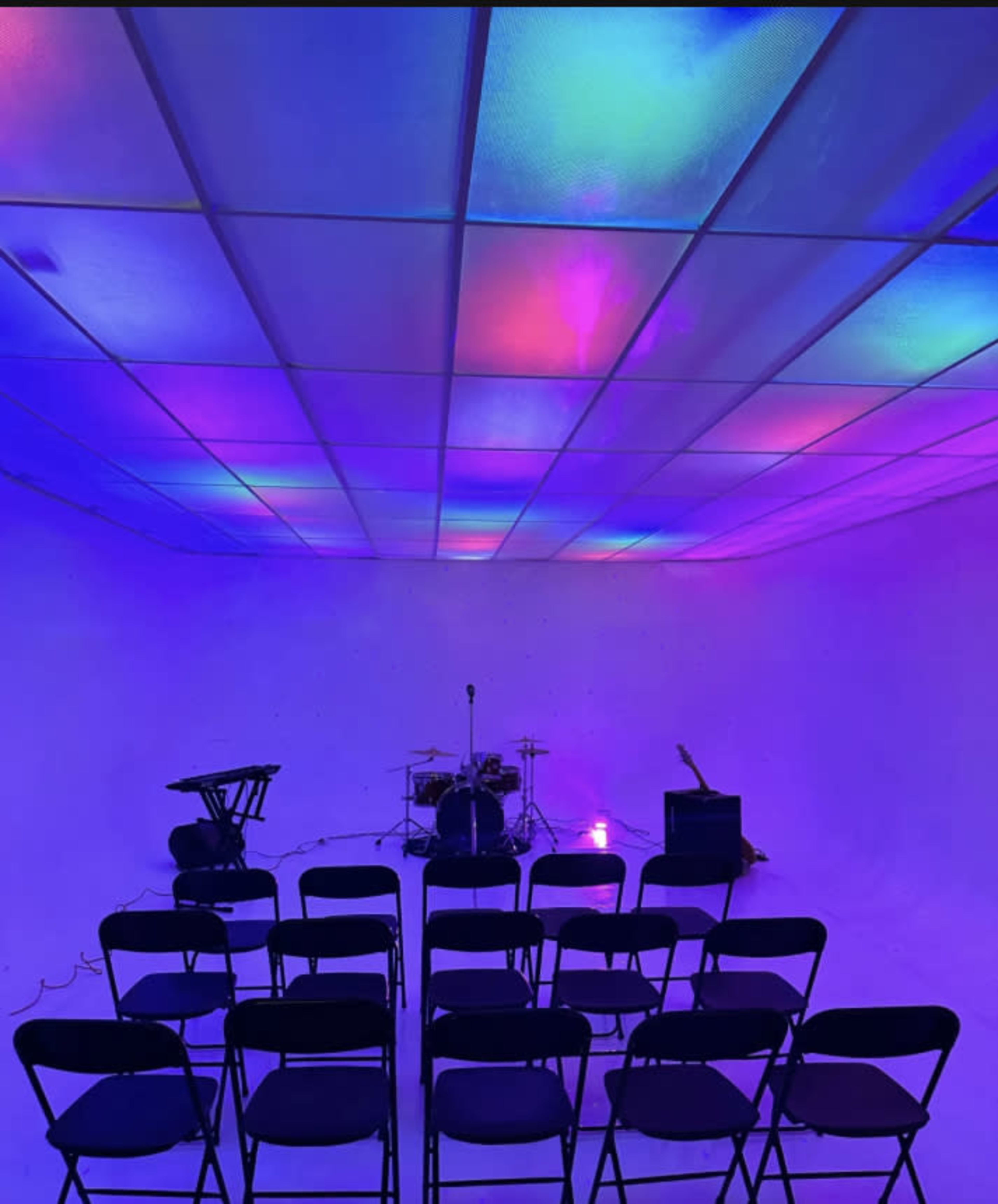 An empty venue with black chairs arranged in rows facing a small stage equipped with musical instruments and colorful lights on the ceiling.