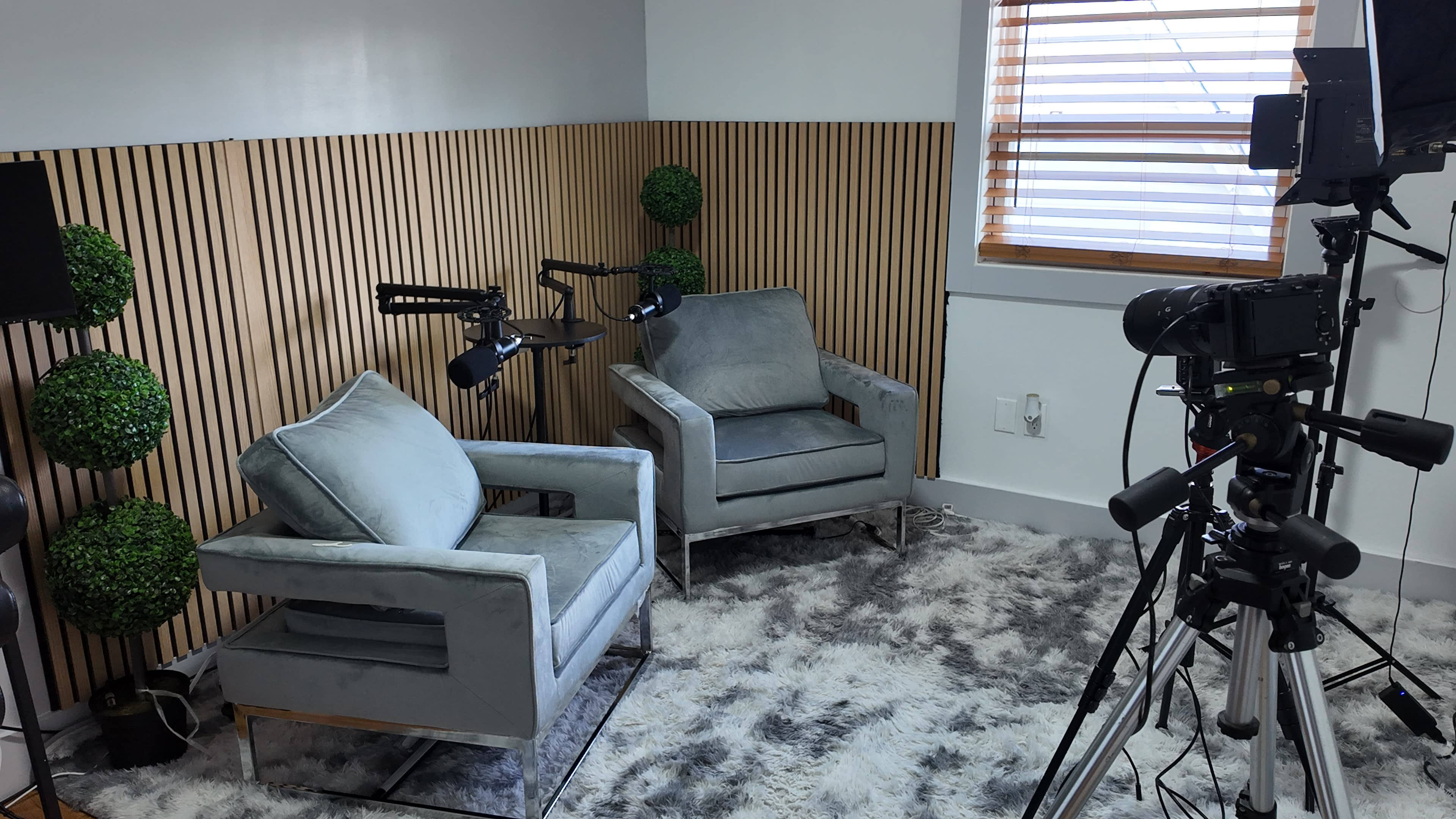 A tidy, modern studio setup featuring two upholstered chairs, a textured rug, and camera equipment positioned for filming.