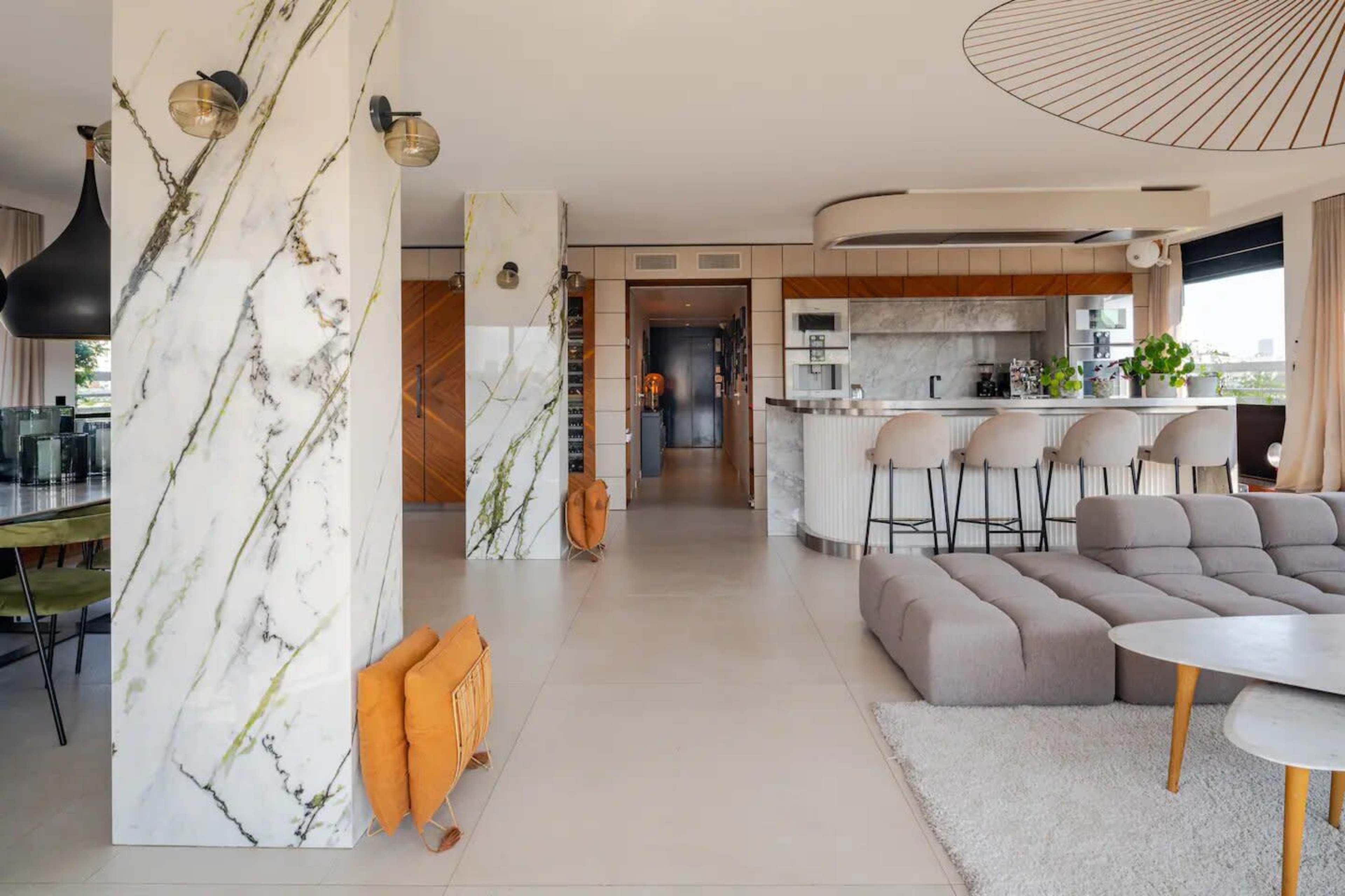 A modern, open-concept living space featuring marble columns, a kitchen island with seating, and a beige sectional sofa.