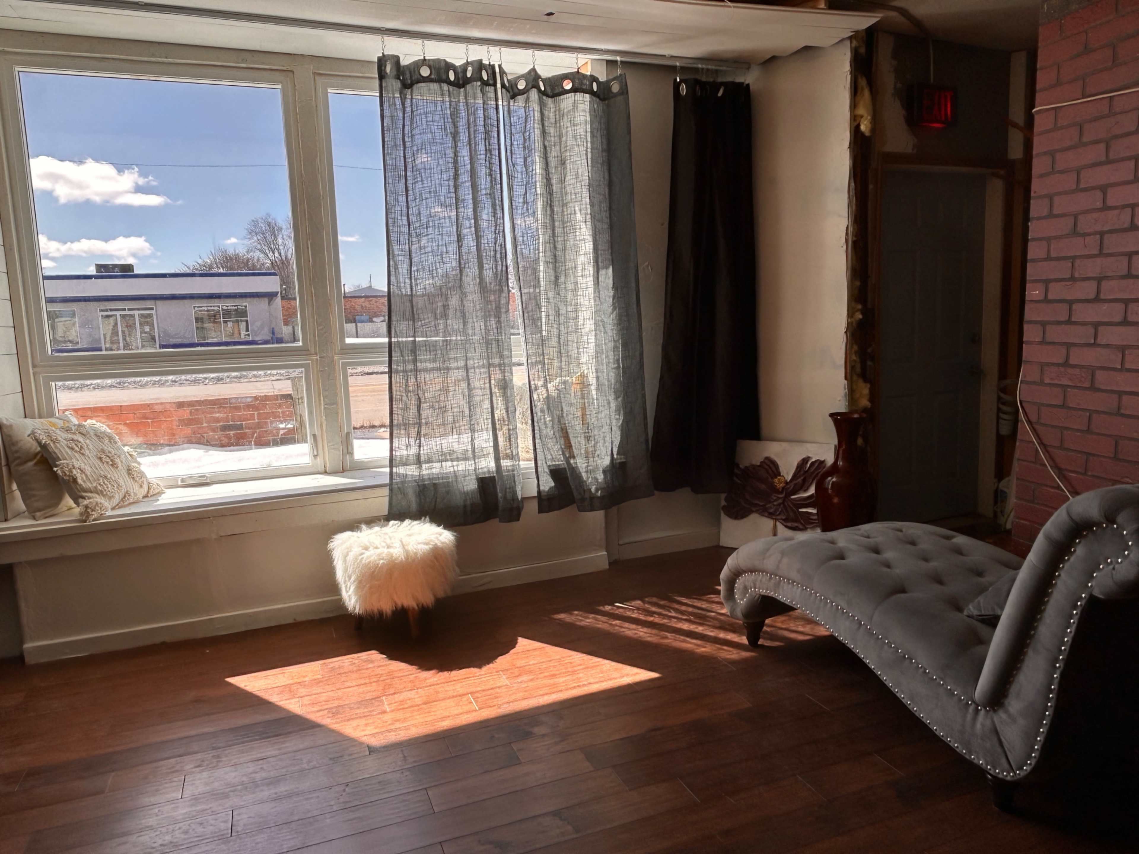 A sunlit corner of a room features a chaise lounge, a fluffy ottoman, and sheer curtains overlooking an outdoor view.
