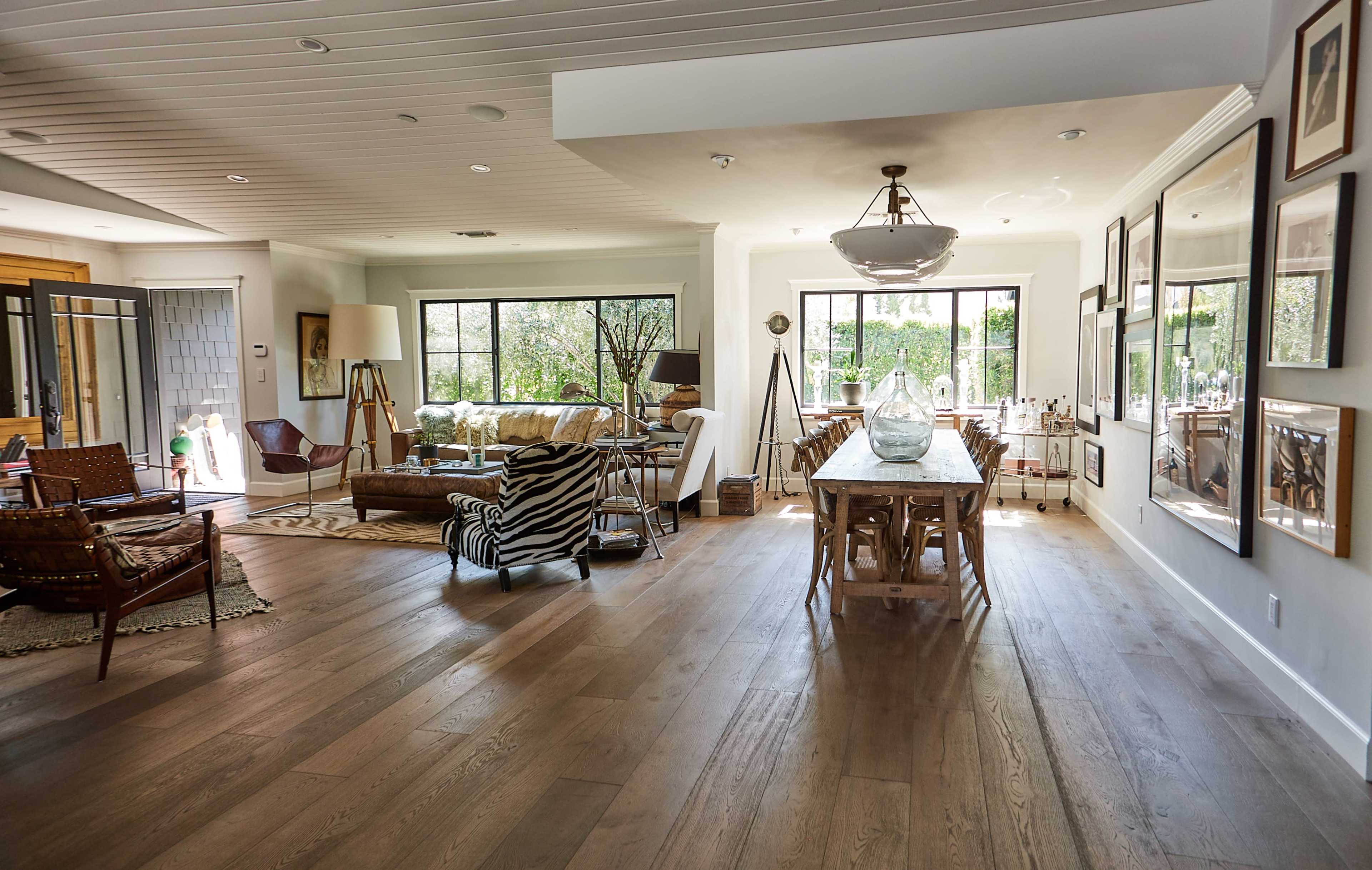 The image depicts a spacious, open-concept living area featuring a dining table, various seating options, and large windows allowing natural light to fill the room.