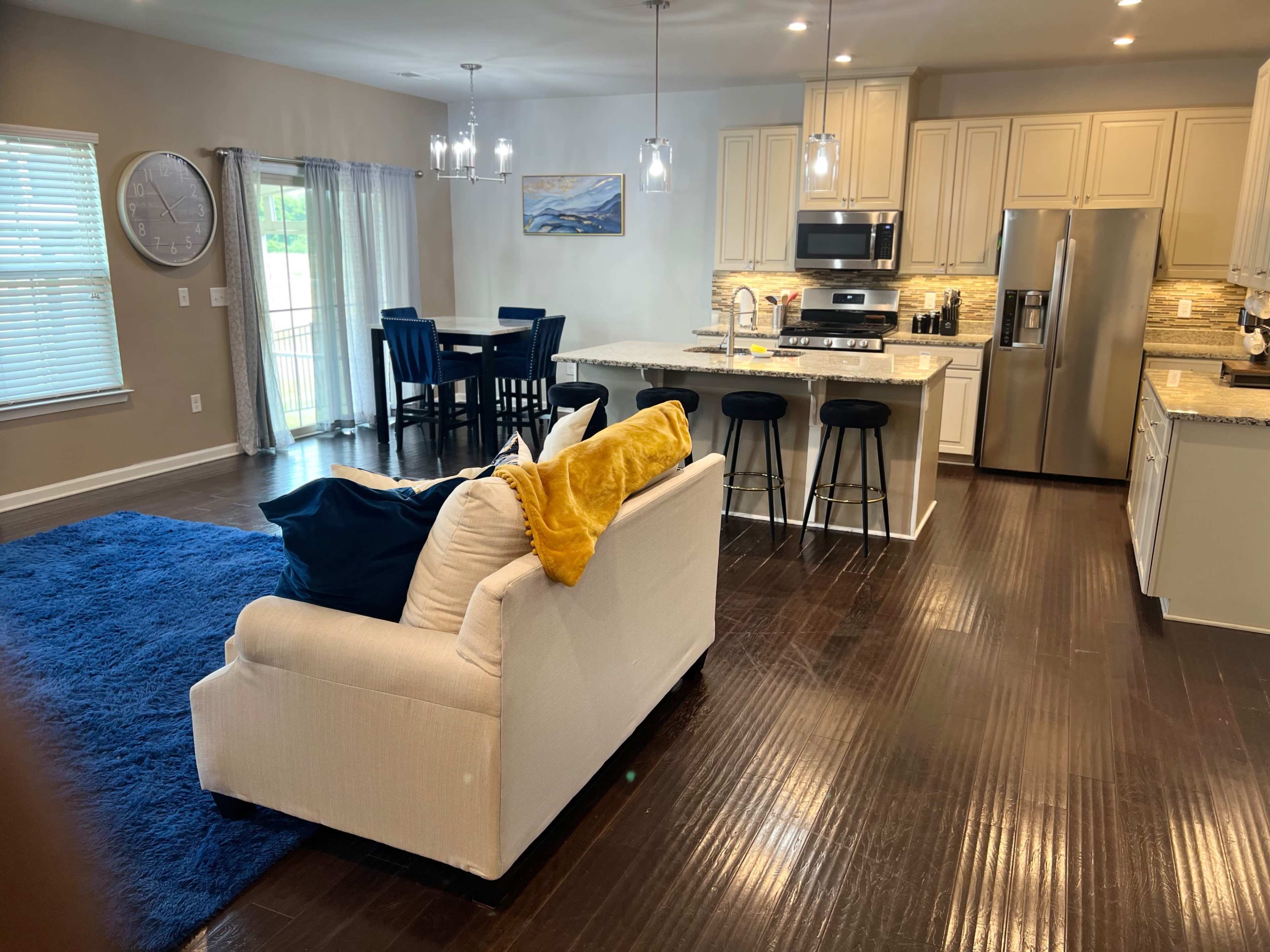 A modern open-concept living area with a white sofa, a blue rug, a kitchen with granite countertops, and a dining table nearby.