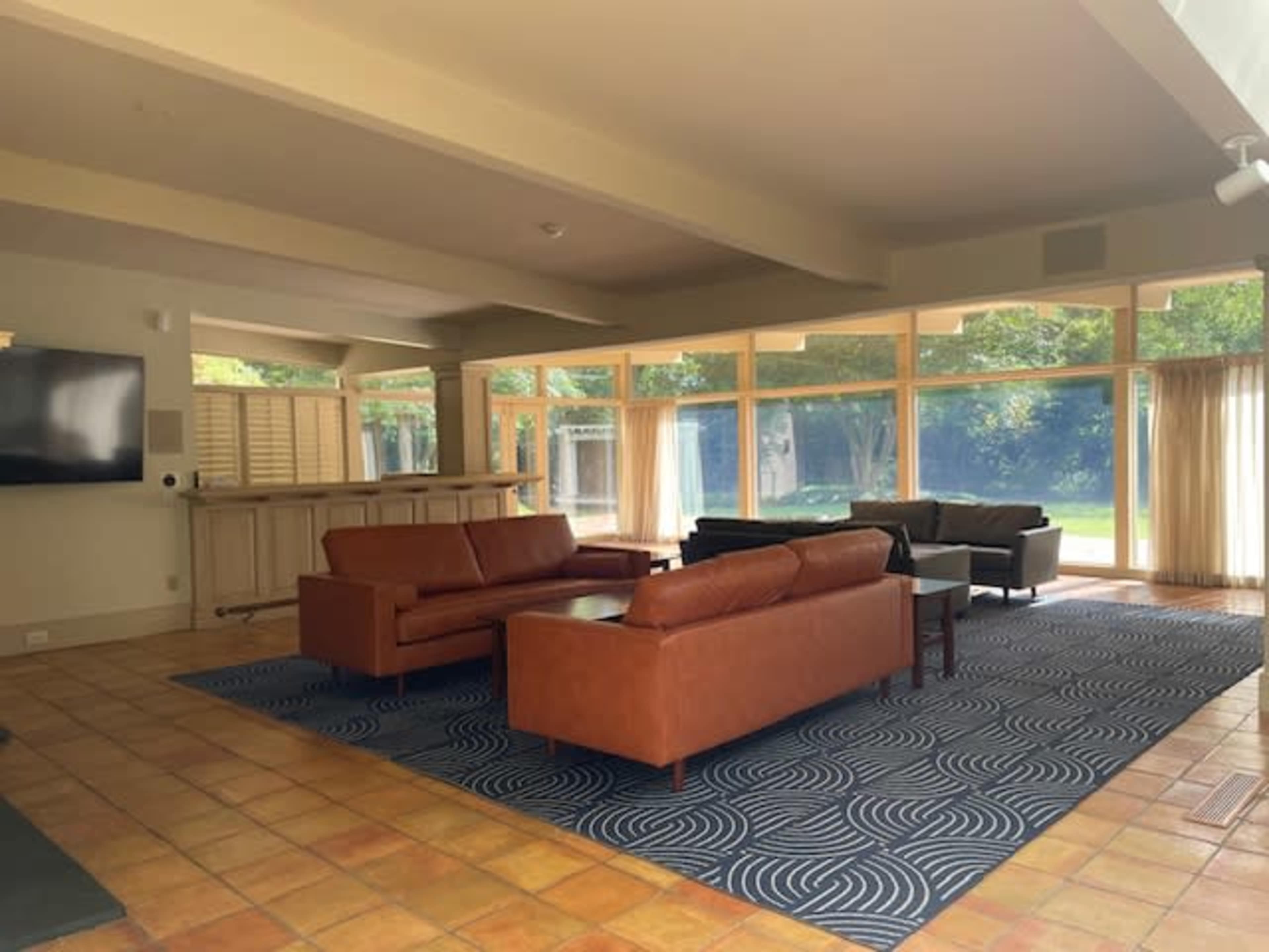 A spacious room with large windows, featuring two brown leather sofas, a black couch, and a patterned area rug.