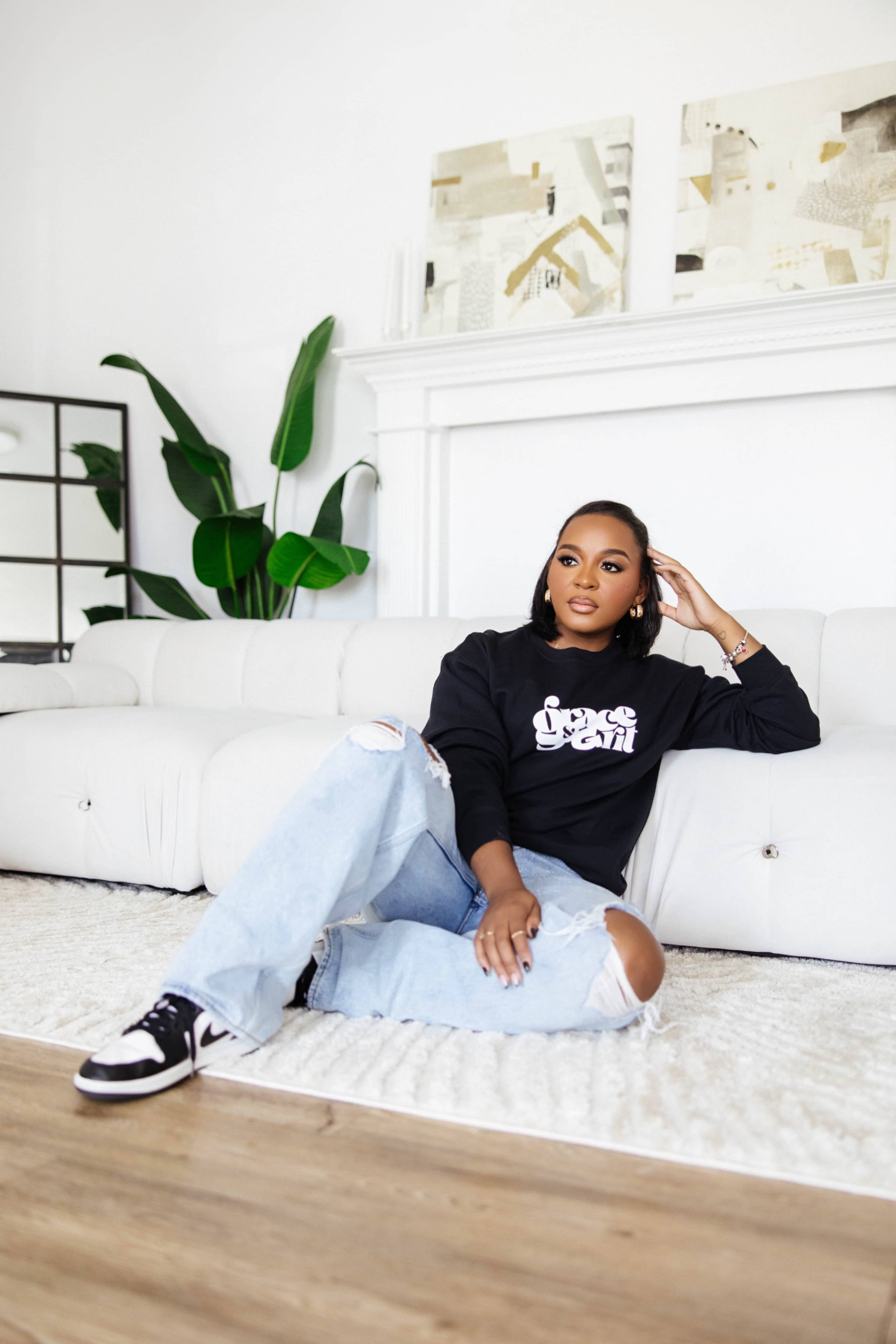 A young woman sits on a rug in front of a white couch, wearing a black sweatshirt and light blue ripped jeans, with a large plant visible in the background.