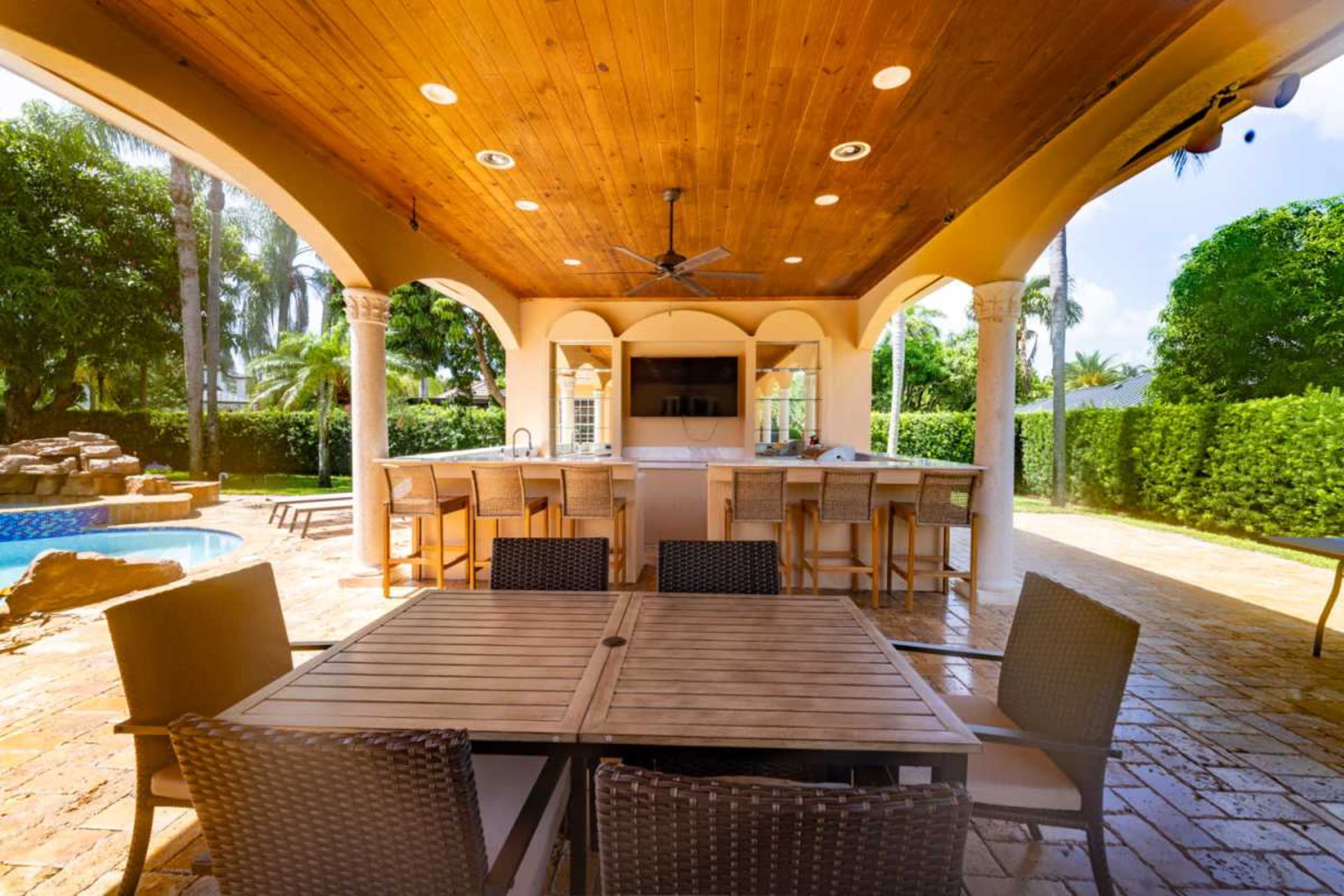 The image shows a shaded outdoor patio area with a large wooden table and chairs, an outdoor kitchen with bar seating, and a swimming pool in the background.