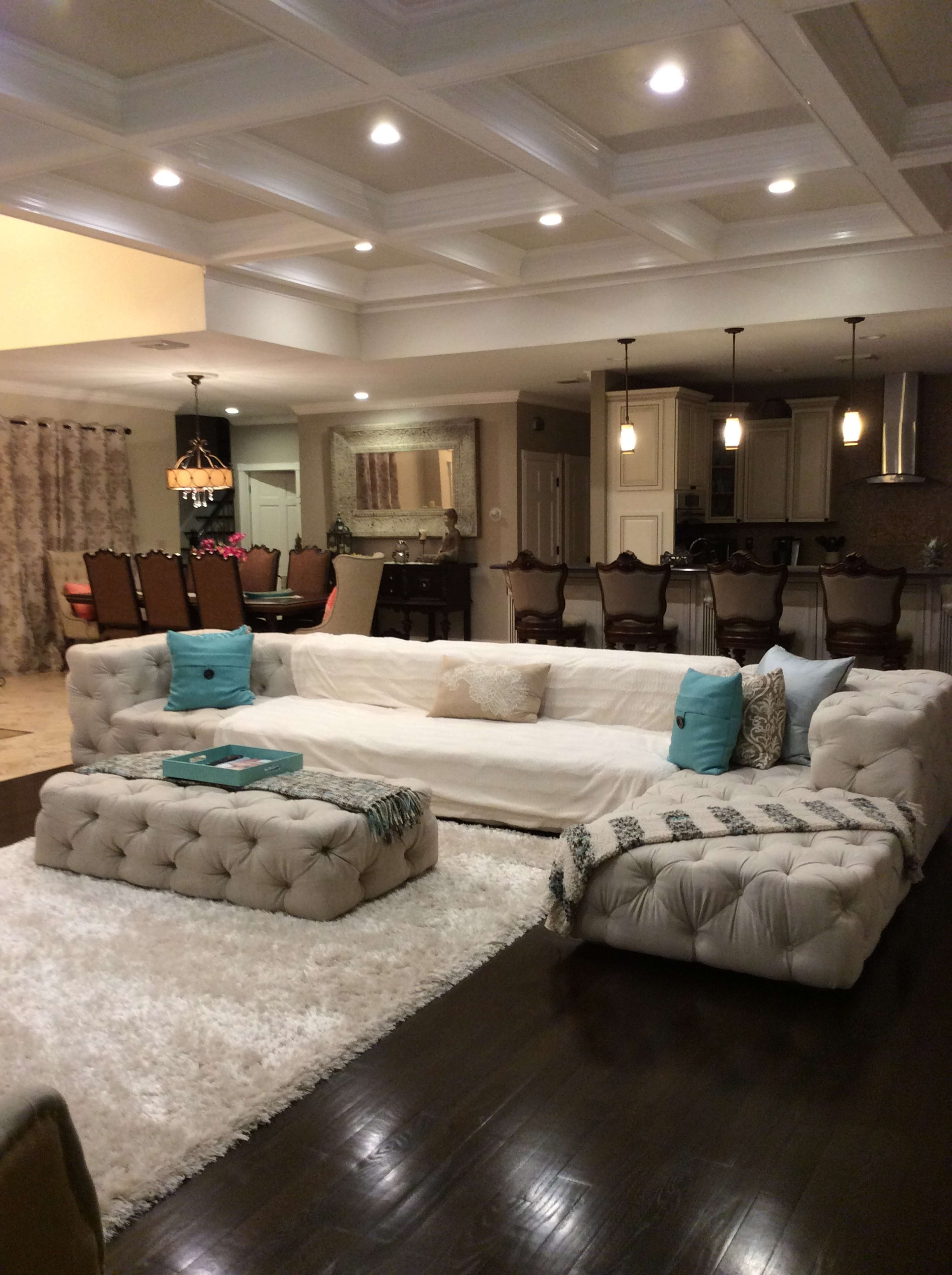 The living room features a large tufted sectional sofa, a plush white area rug, and an open kitchen area with barstools in the background.