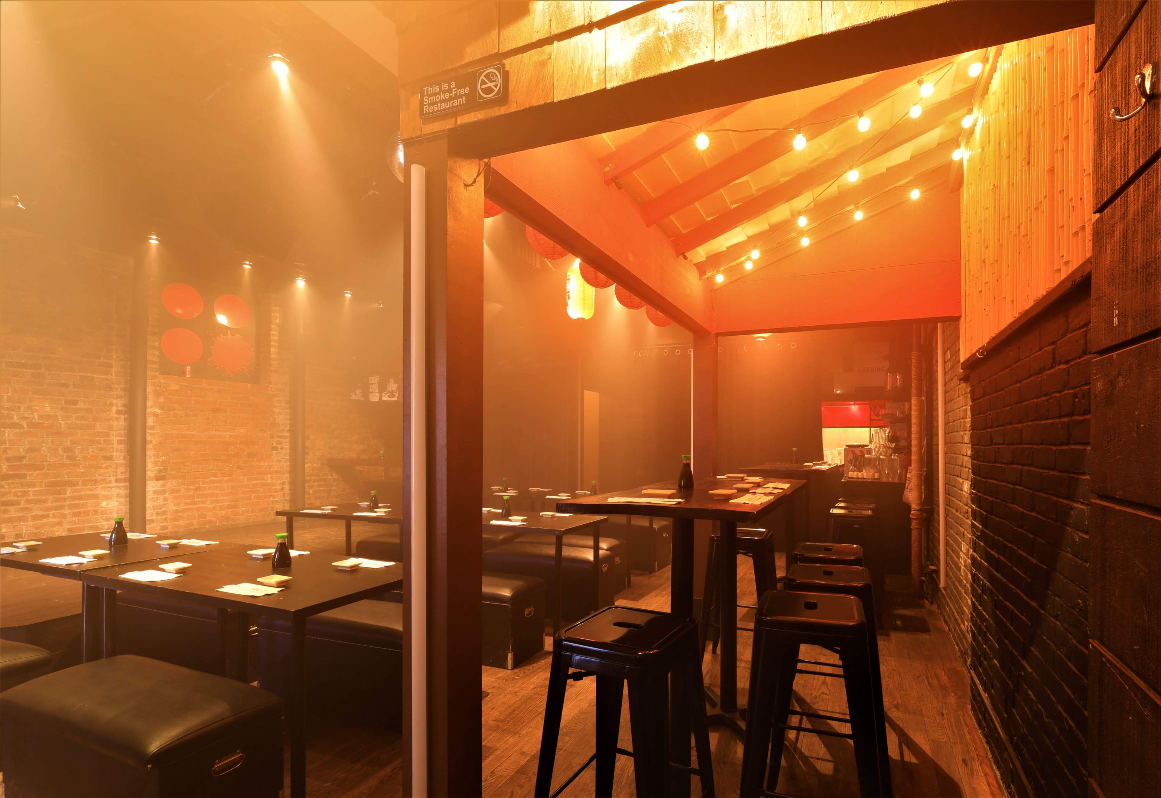 The image shows an empty restaurant interior with wooden seating, exposed brick walls, and soft lighting from hanging bulbs.