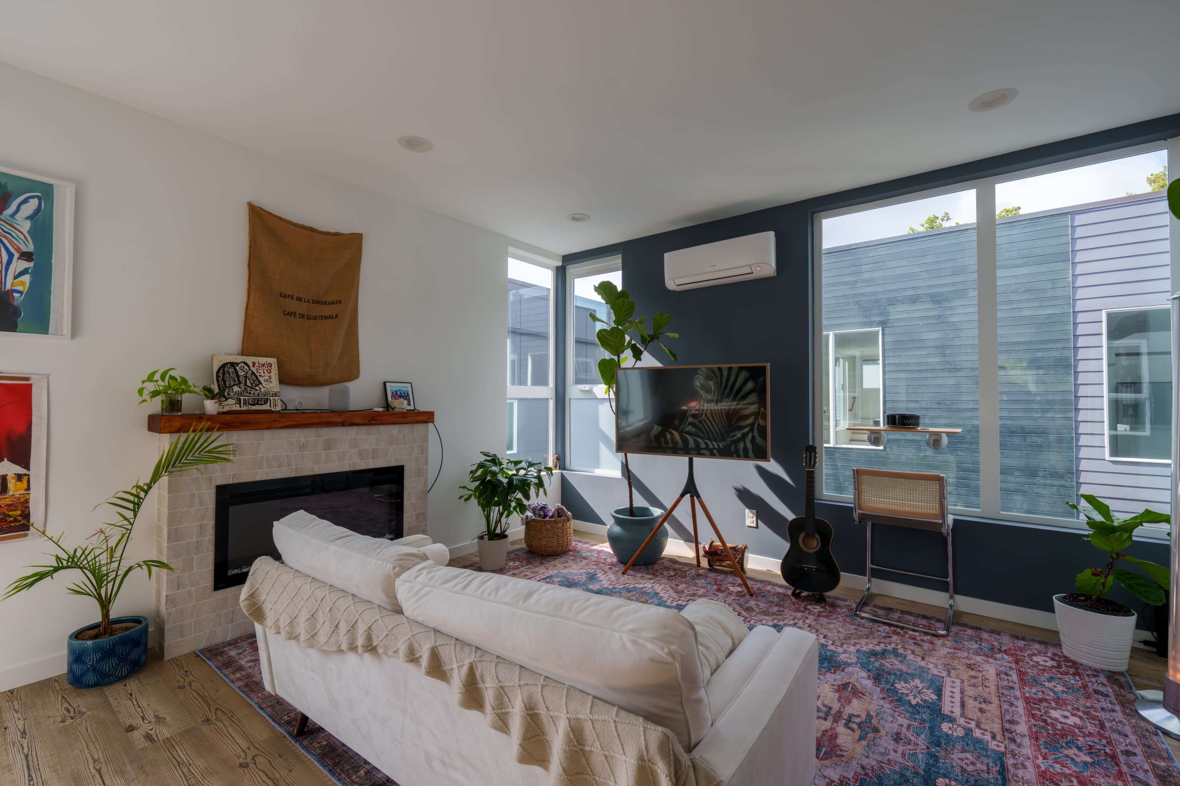 A modern living room features a white couch, a fireplace, a large television mounted on a wall, and indoor plants, with large windows allowing natural light.