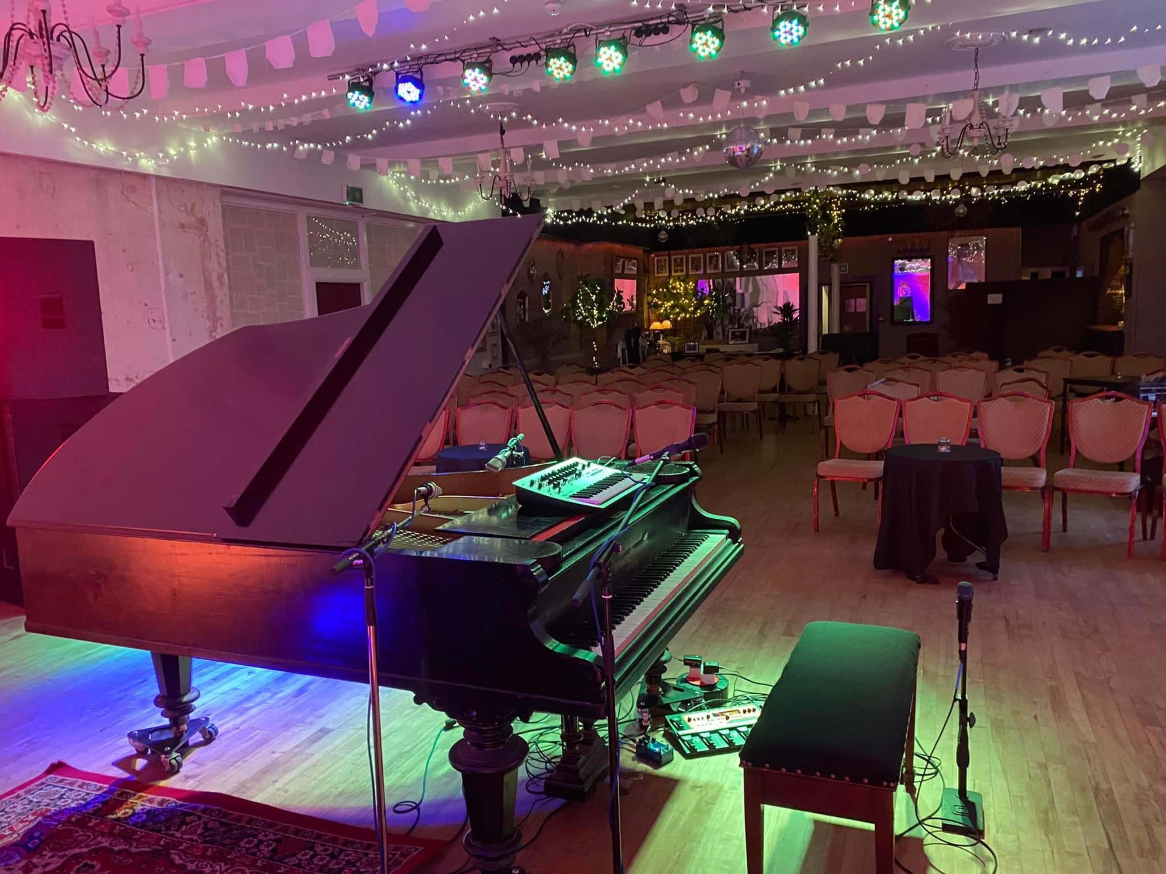 A grand piano is positioned in a well-lit room set up for an event, with chairs arranged in rows and decorative lights hanging from the ceiling.
