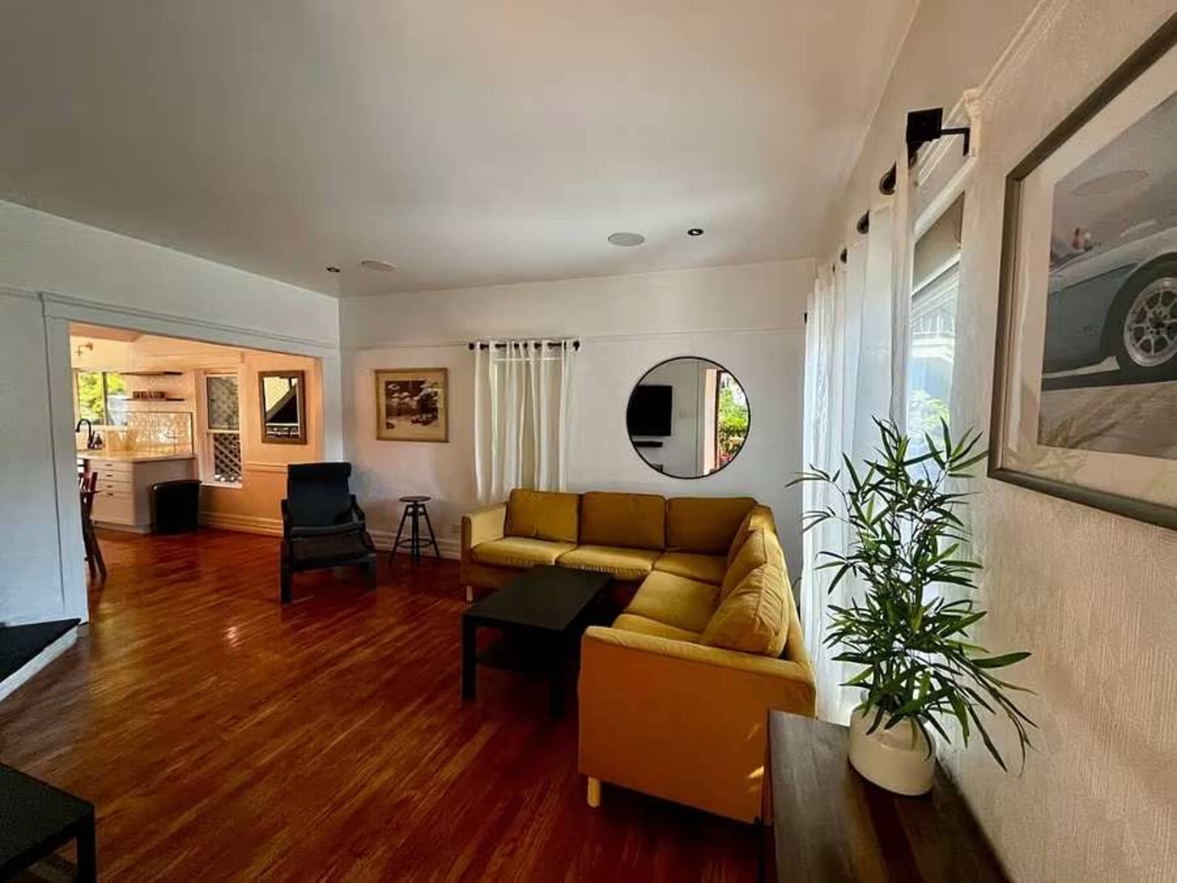 The image shows a bright, open living room with a yellow sectional sofa, a black coffee table, and a plant, leading to a kitchen area in the background.