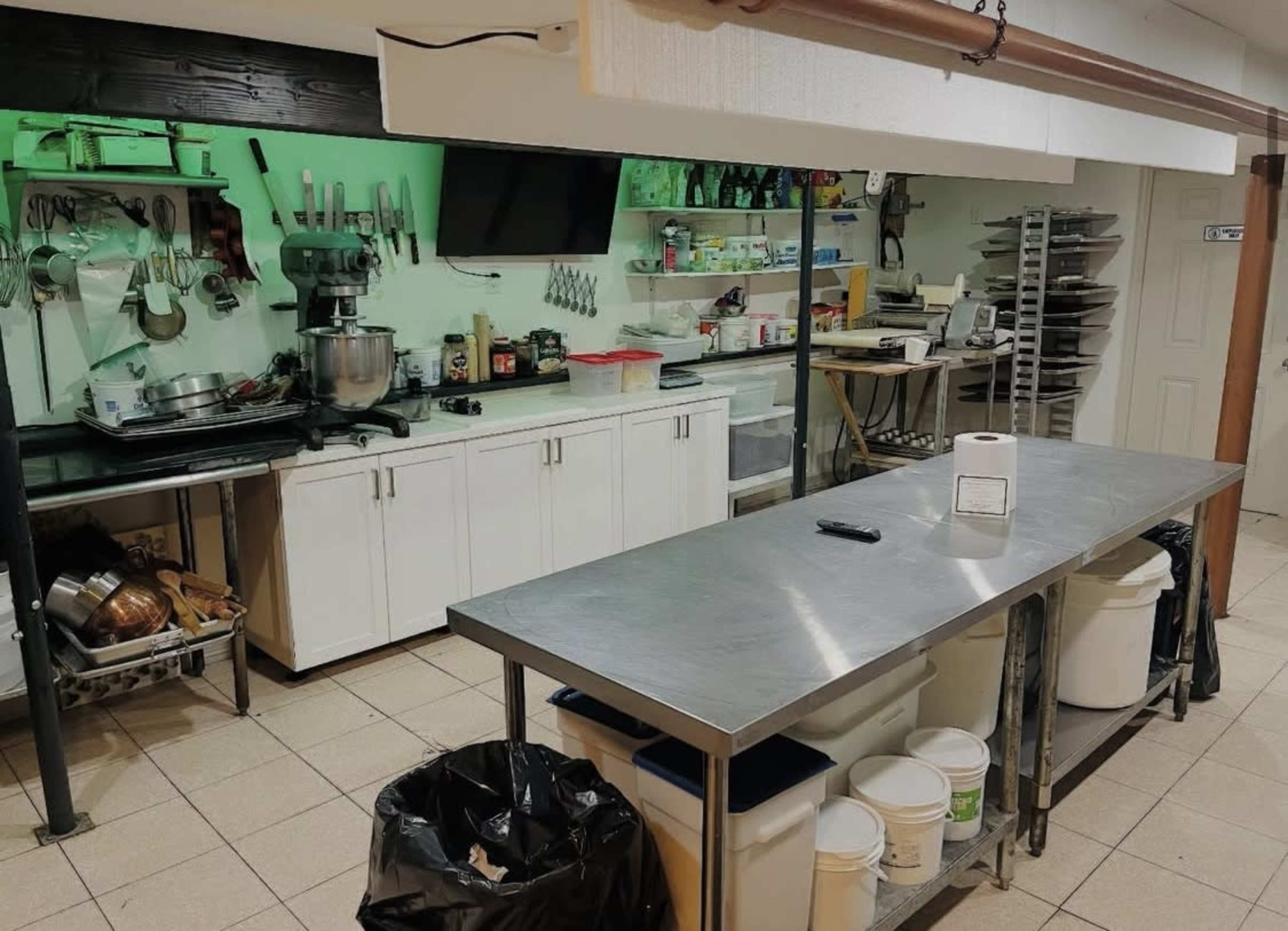 The image shows a kitchen with stainless steel countertops, various cooking utensils on the walls, and storage shelves filled with food items and equipment.
