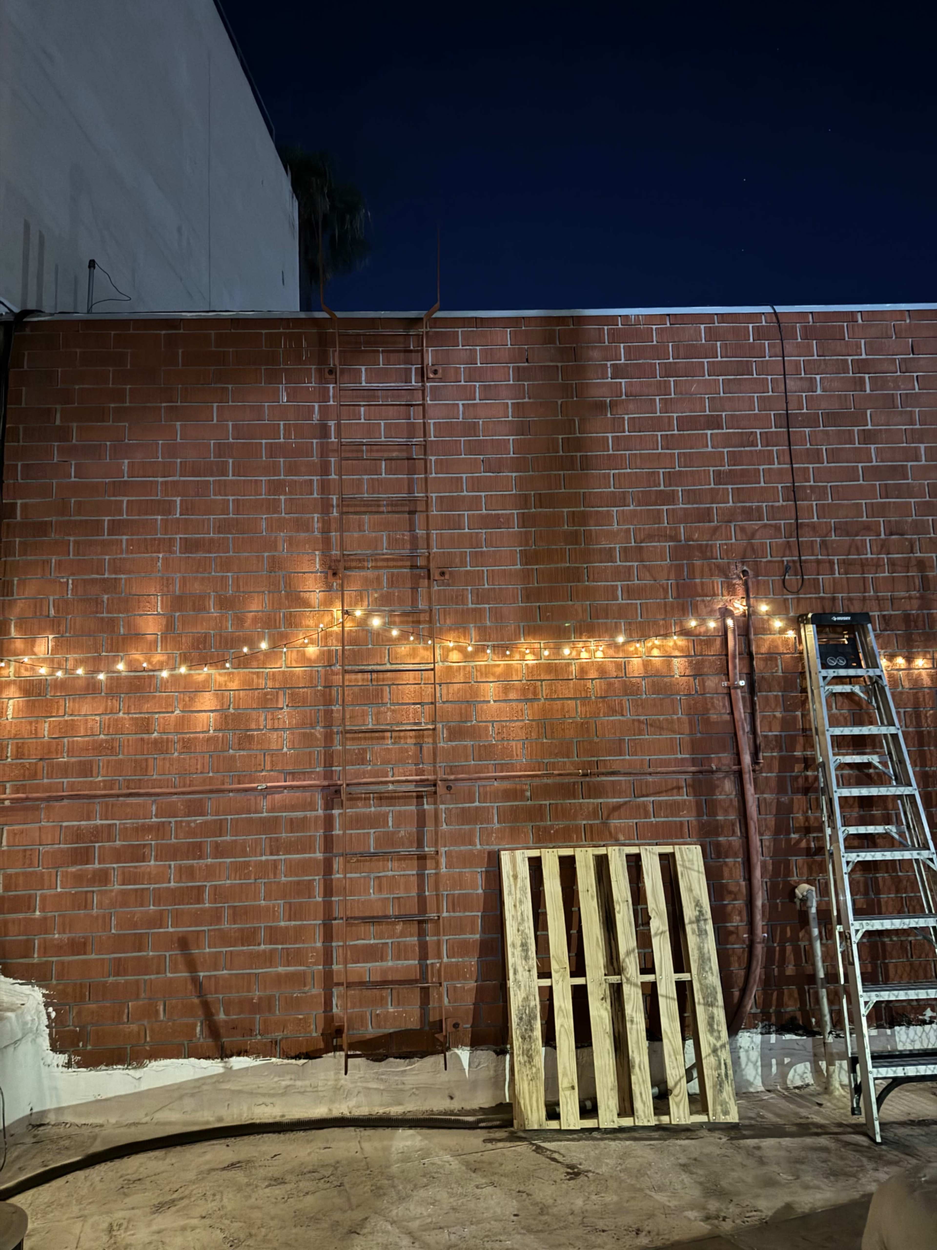 The image shows a brick wall at night, illuminated by string lights, with a wooden pallet and a ladder leaning against it.