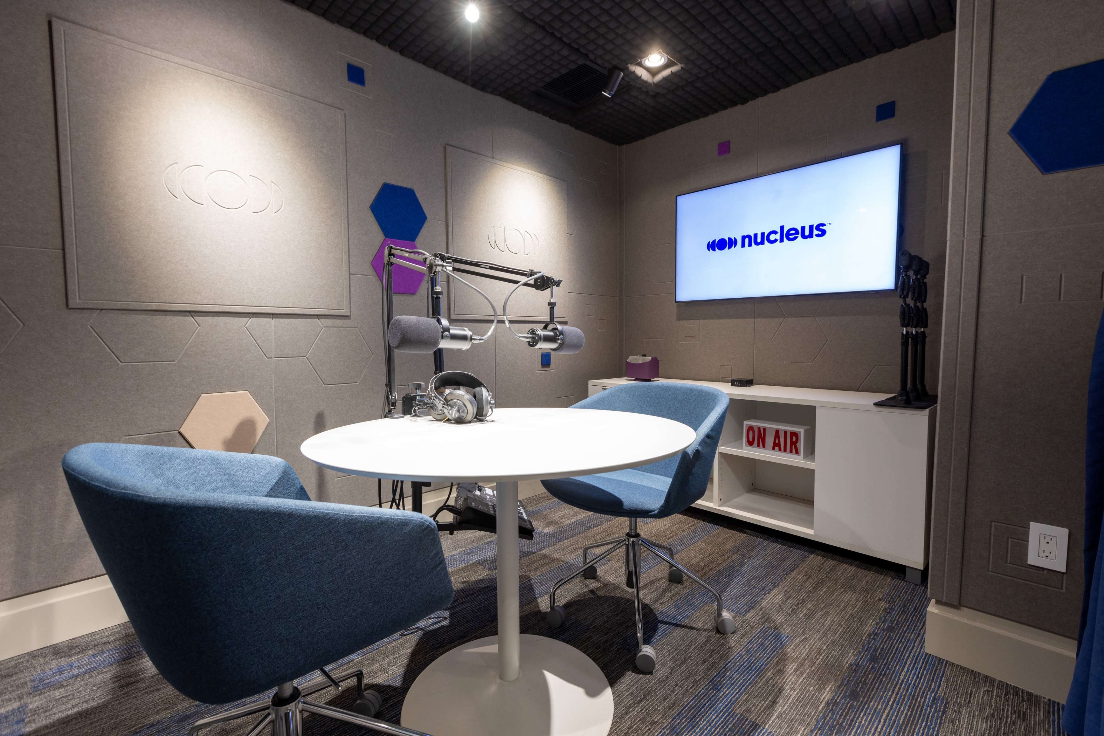 A small, modern recording studio featuring two blue chairs, a round table, a large screen displaying "nucleus," and a microphone setup against a soundproof wall.