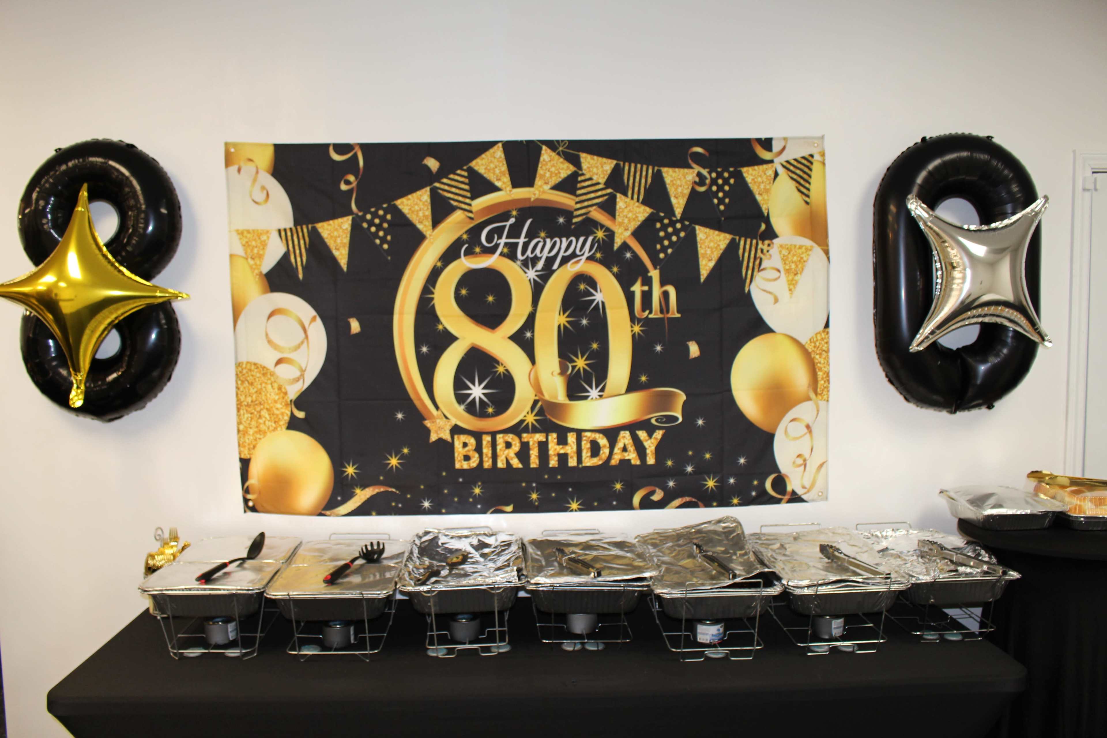 A table is set up for a birthday celebration featuring a large banner that reads "Happy 80th Birthday" along with gold and black decorations, and several covered food trays.