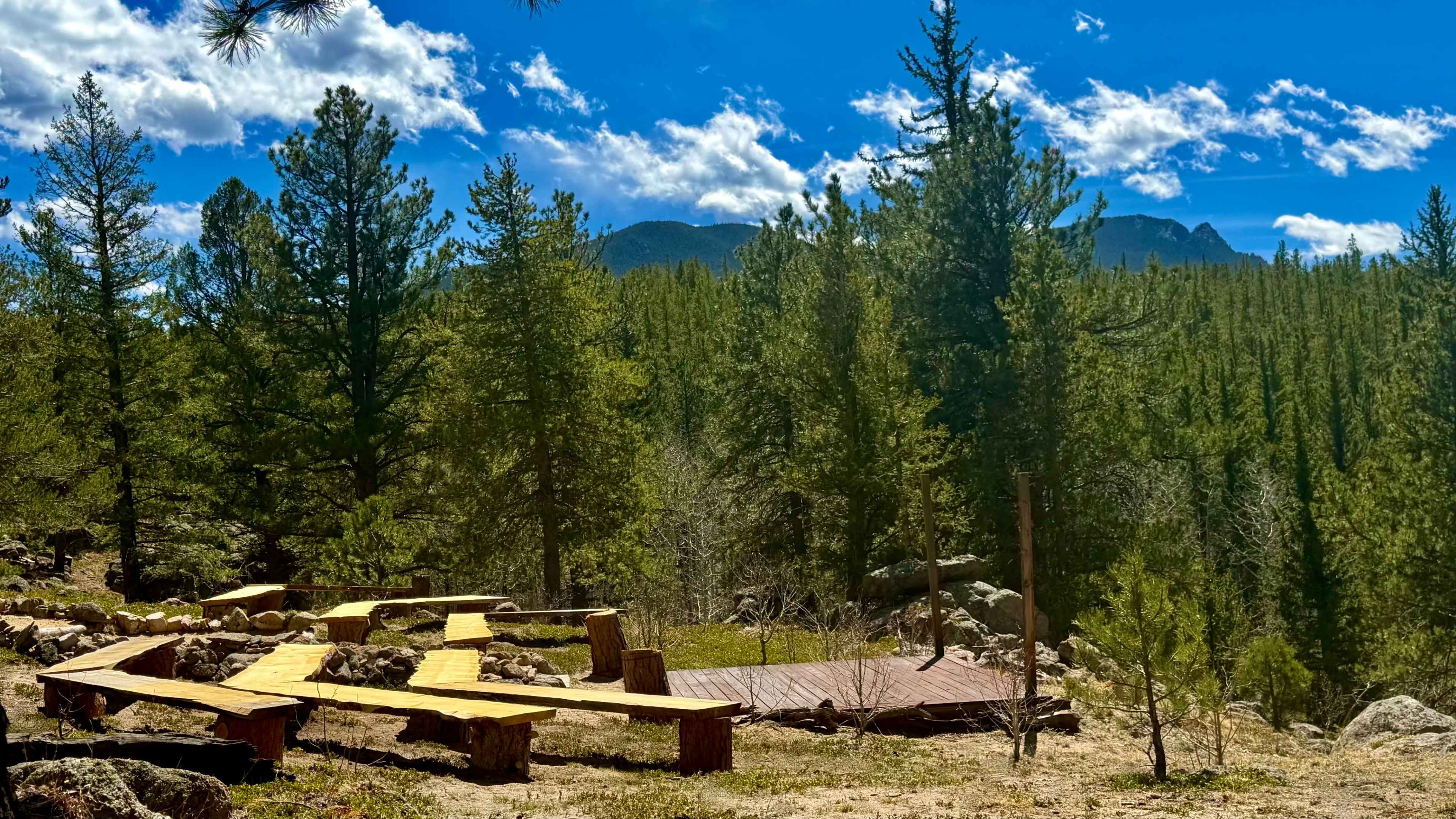 A clearing in a forest with wooden benches arranged around a designated gathering area, surrounded by tall pine trees and distant mountain peaks under a blue sky.