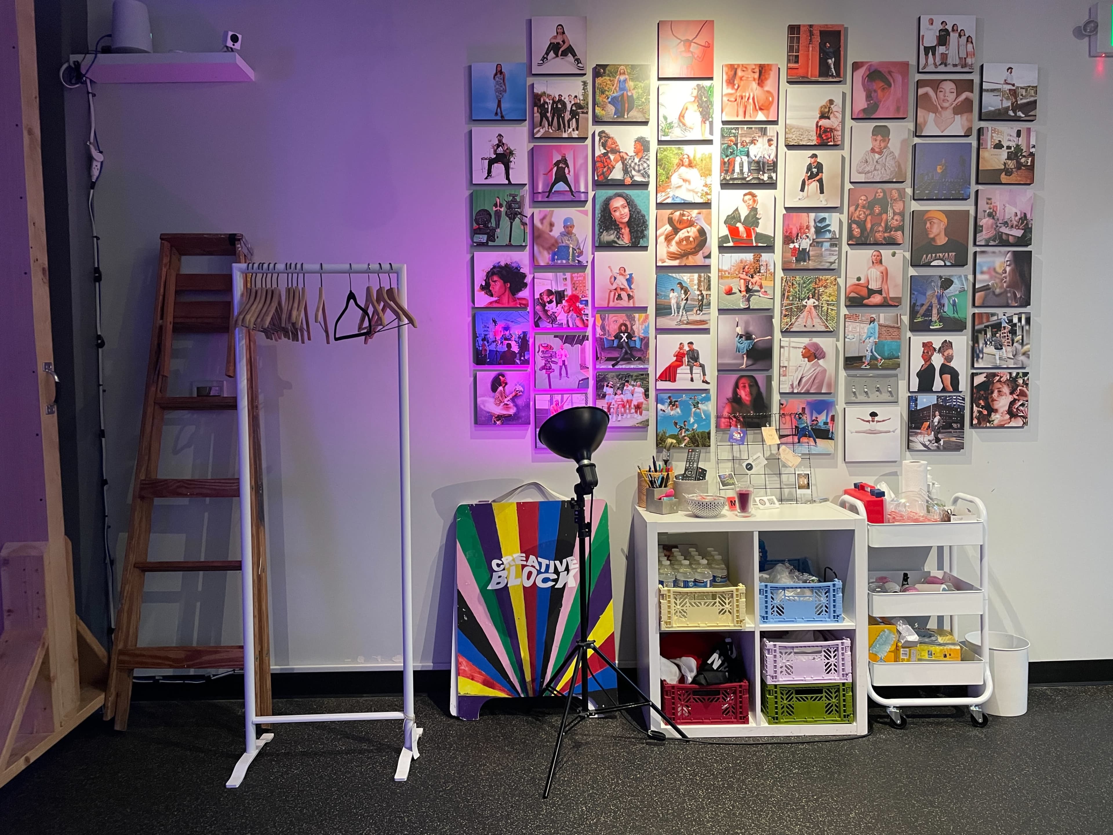 A colorful studio space with a wall covered in framed photographs, a clothing rack with hangers, a chair, and a storage unit filled with various containers.