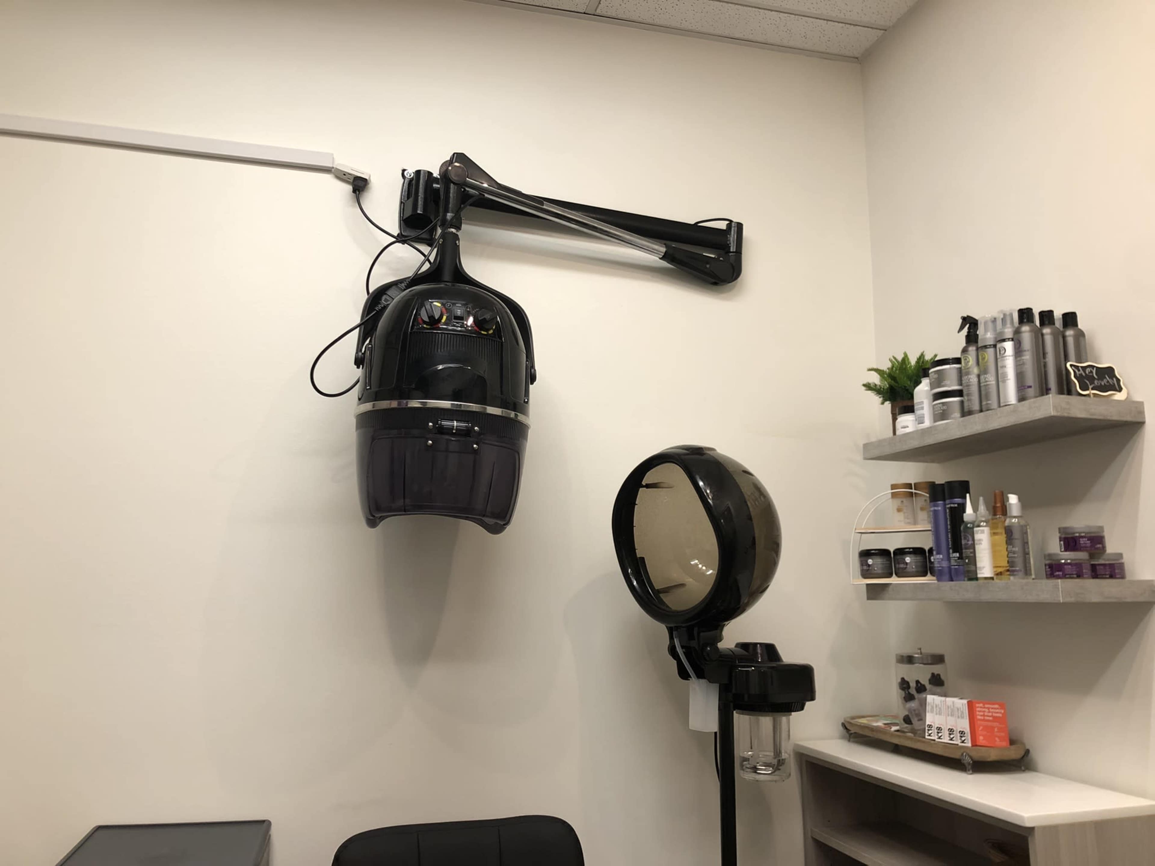 A hair salon setup featuring a wall-mounted hooded dryer and a standing mirror, with shelves displaying various hair products.