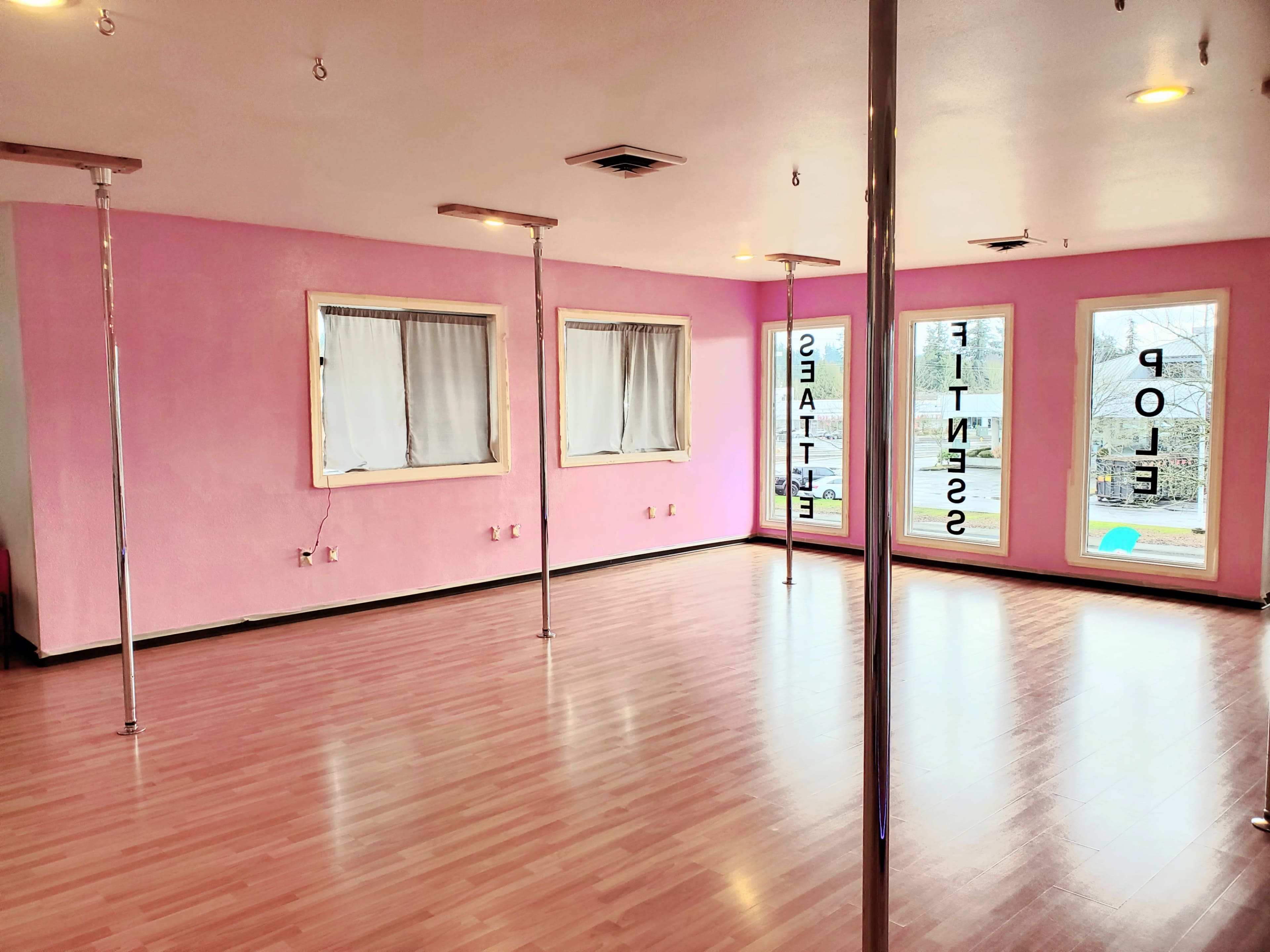 A brightly lit fitness studio with pink walls, polished wooden flooring, and multiple vertical poles.