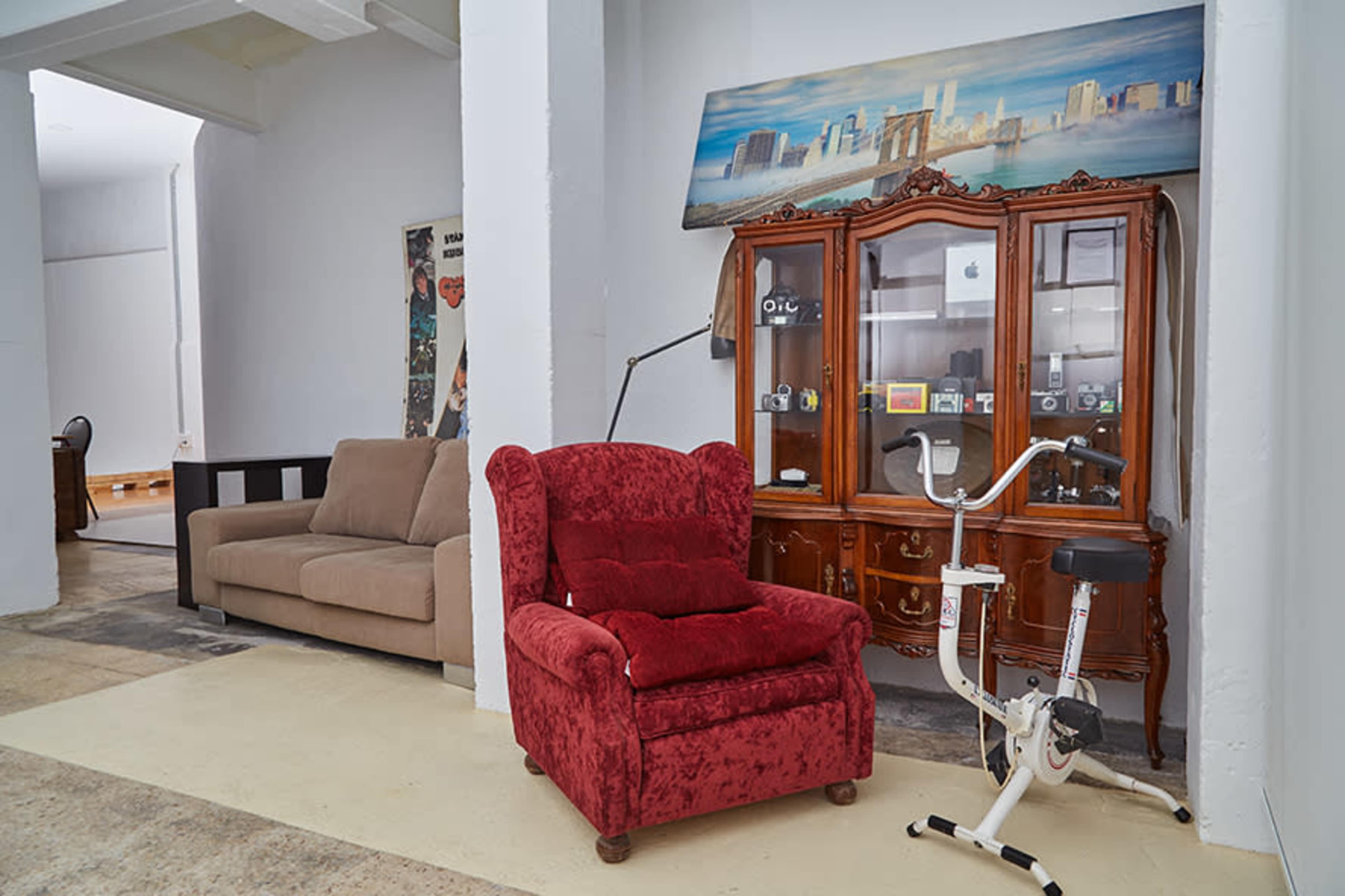 The image features a red upholstered armchair and an exercise bike in a room with a wooden cabinet and a beige couch visible in the background.
