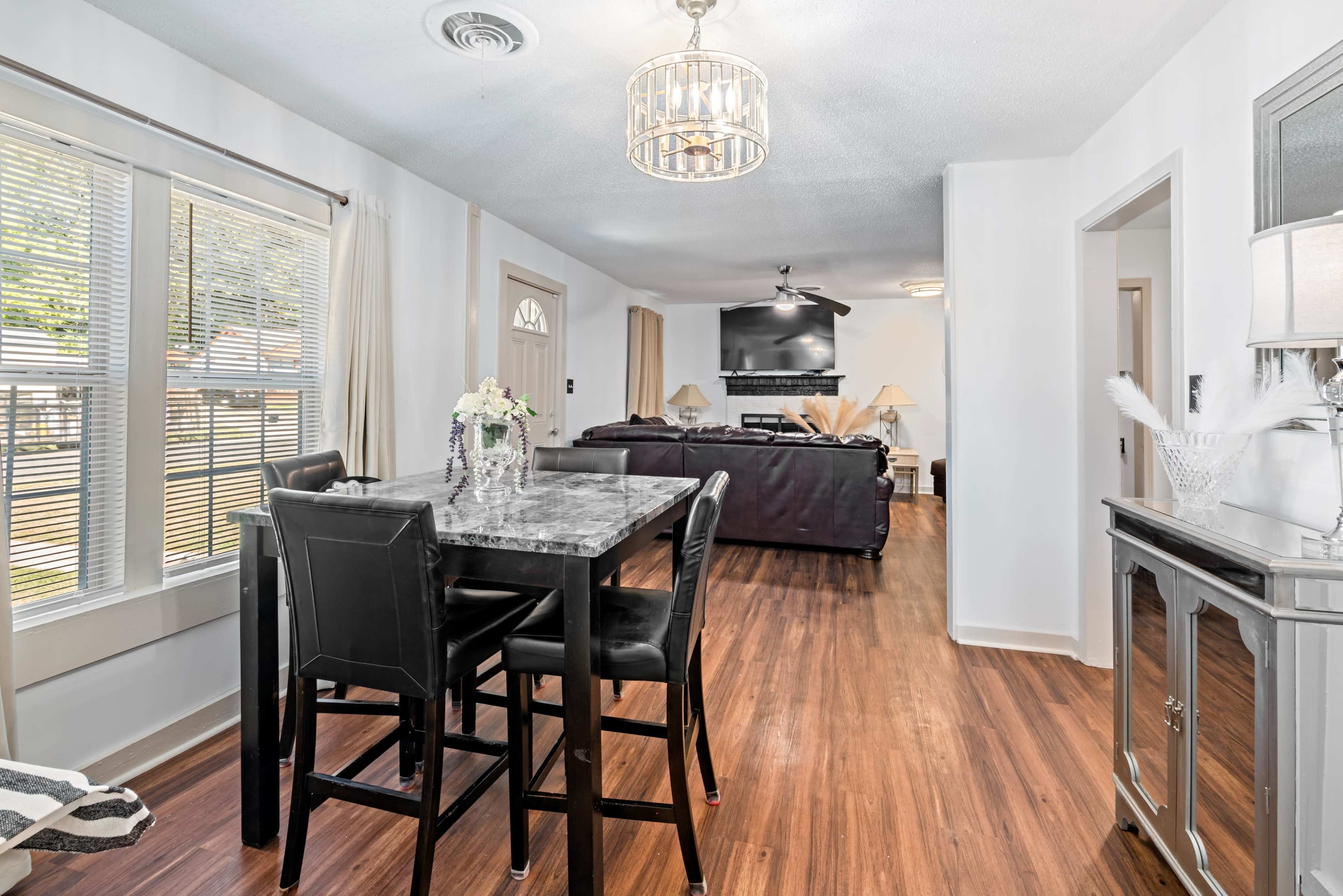 The image shows a modern dining area with a black table and chairs, leading into a living room with a television and seating.