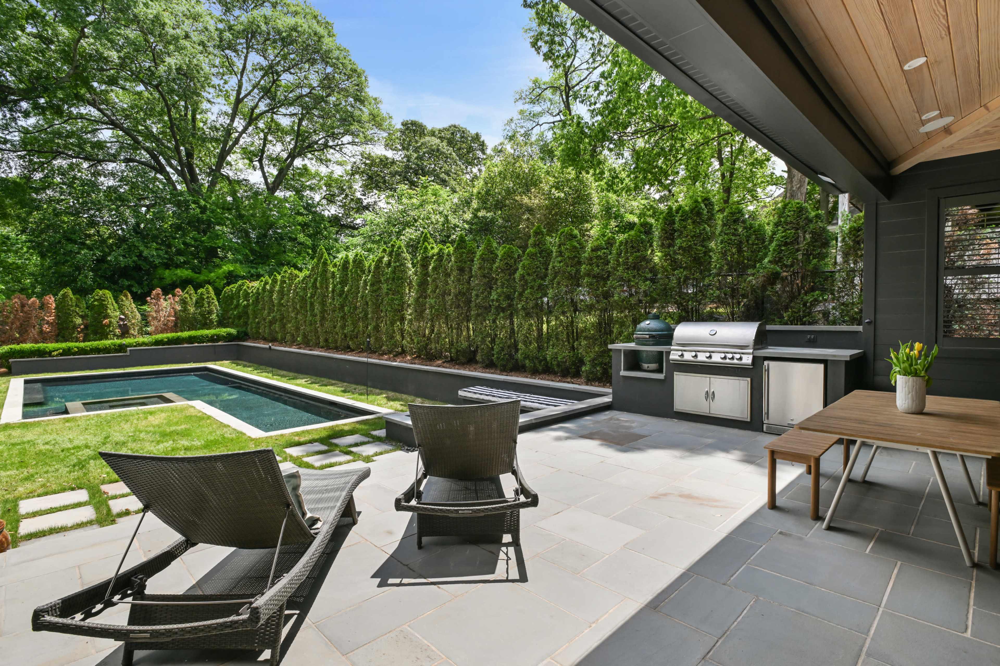The image shows a backyard featuring a swimming pool, outdoor kitchen, and lounge chairs surrounded by neatly trimmed hedges and a patio area.