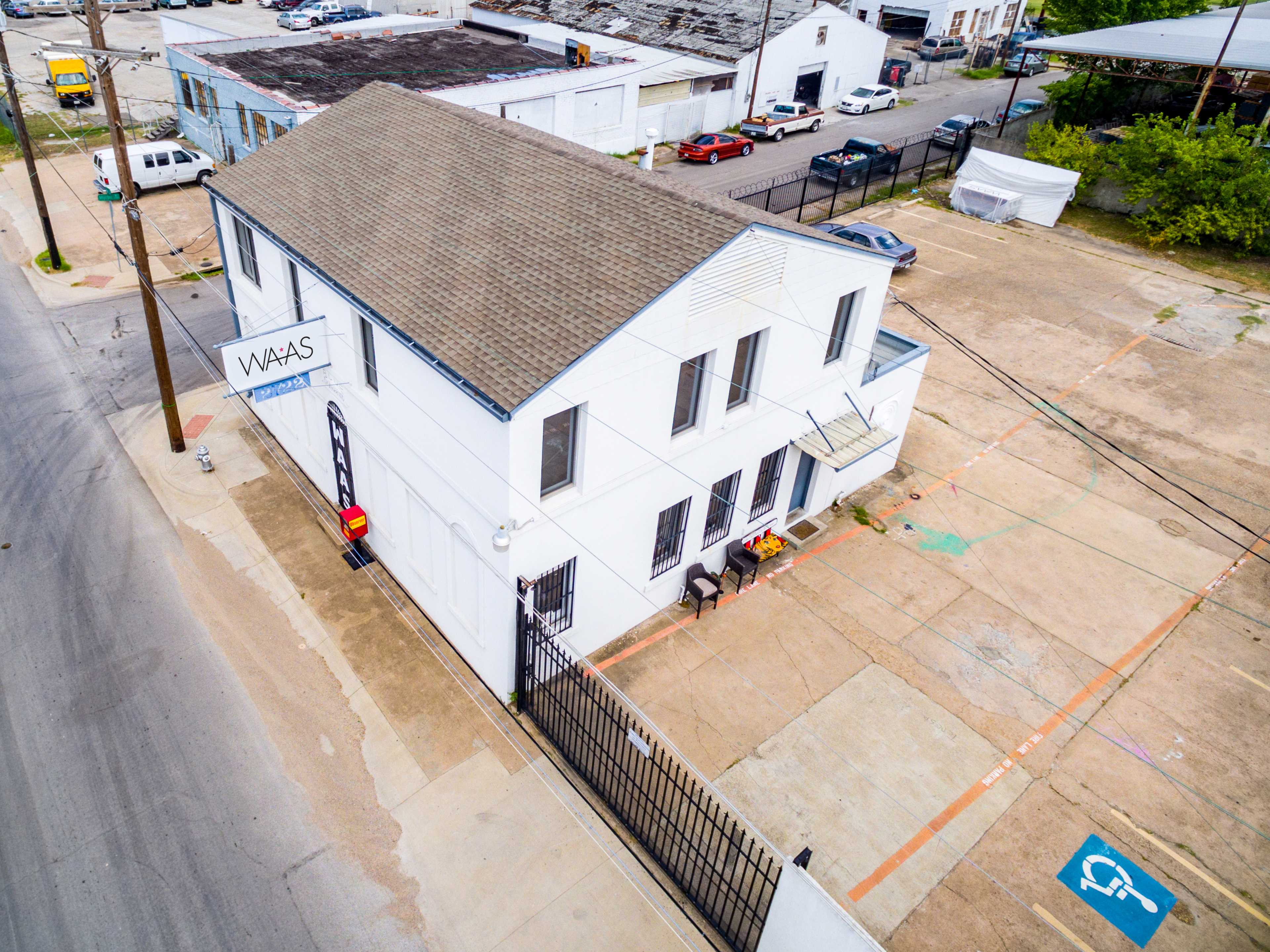 The image shows a two-story white building with a flat roof, surrounded by a large, paved lot and several parked vehicles nearby.
