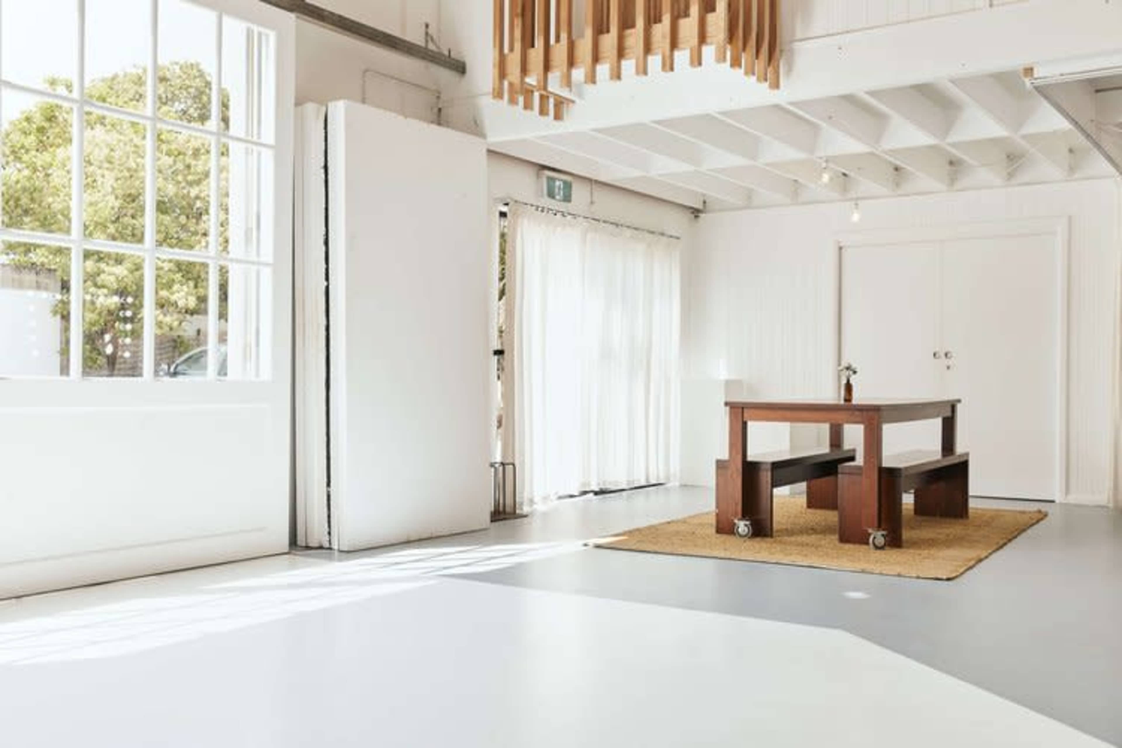 The image shows a bright, minimalist interior with a wooden table on wheels, surrounded by light-colored walls and a large window letting in natural light.