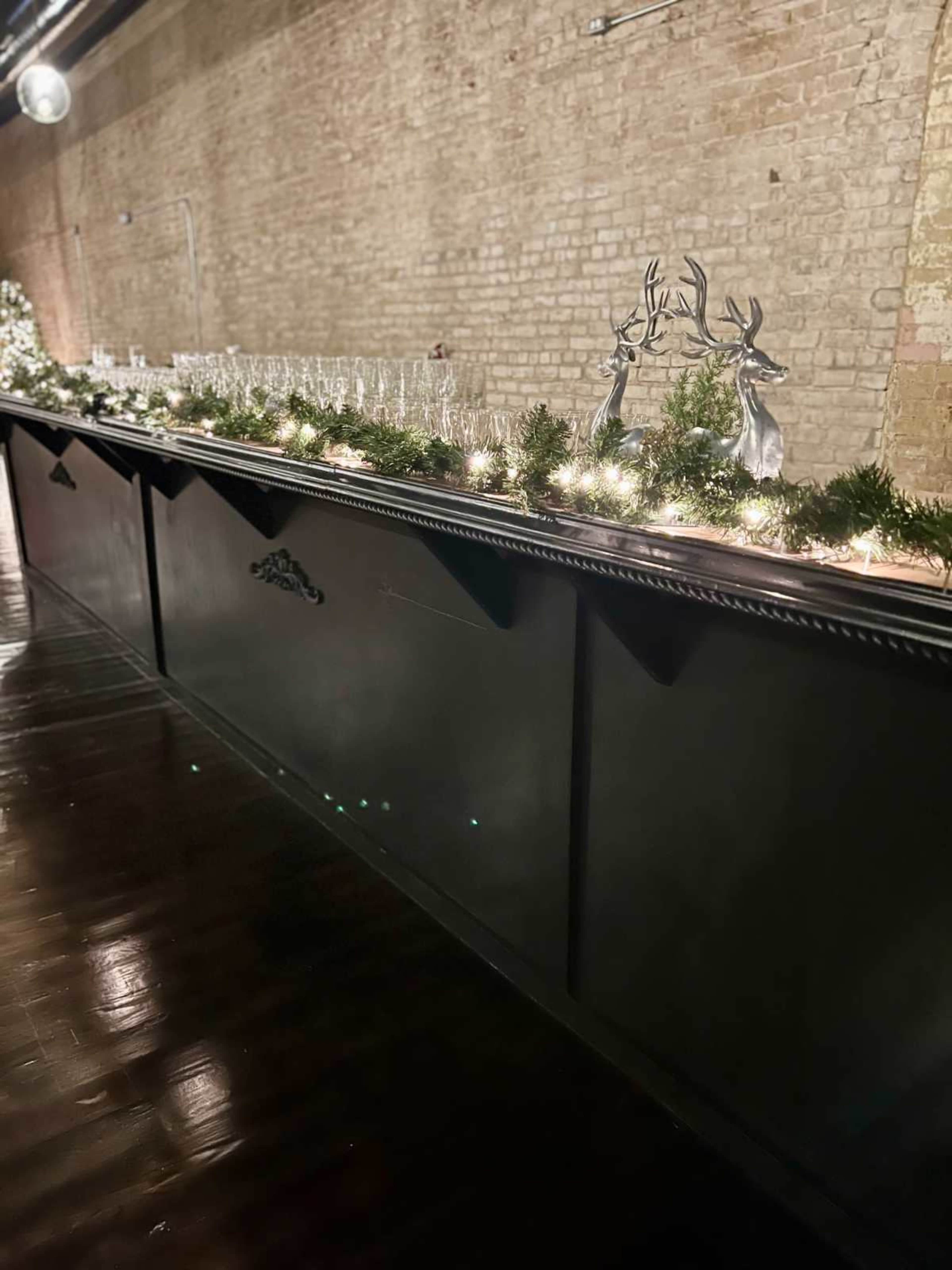A long dark bar adorned with garlands, fairy lights, and decorative silver reindeer against a brick wall backdrop.
