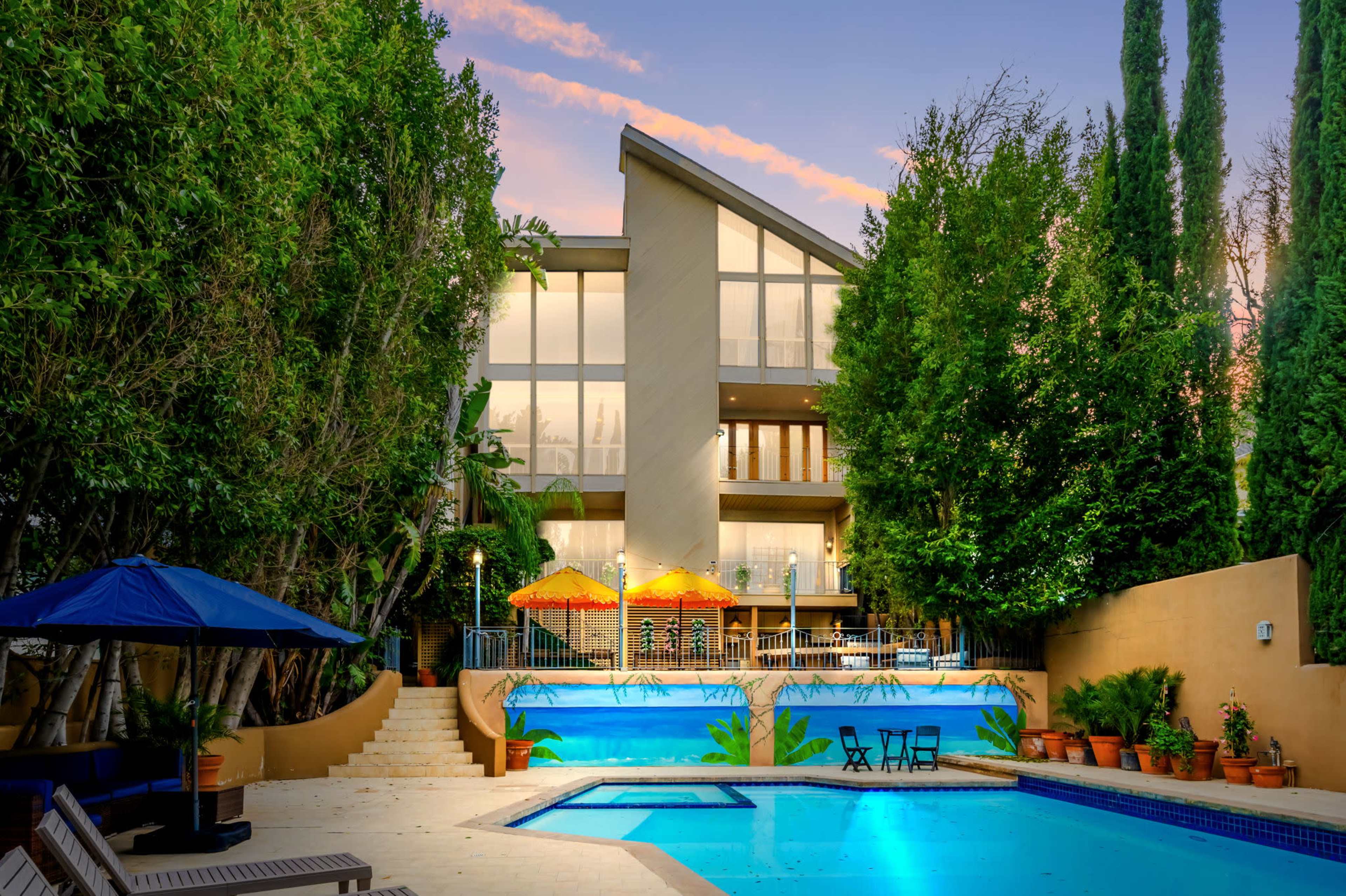 A modern house with large windows overlooks two swimming pools surrounded by greenery and lounge chairs.