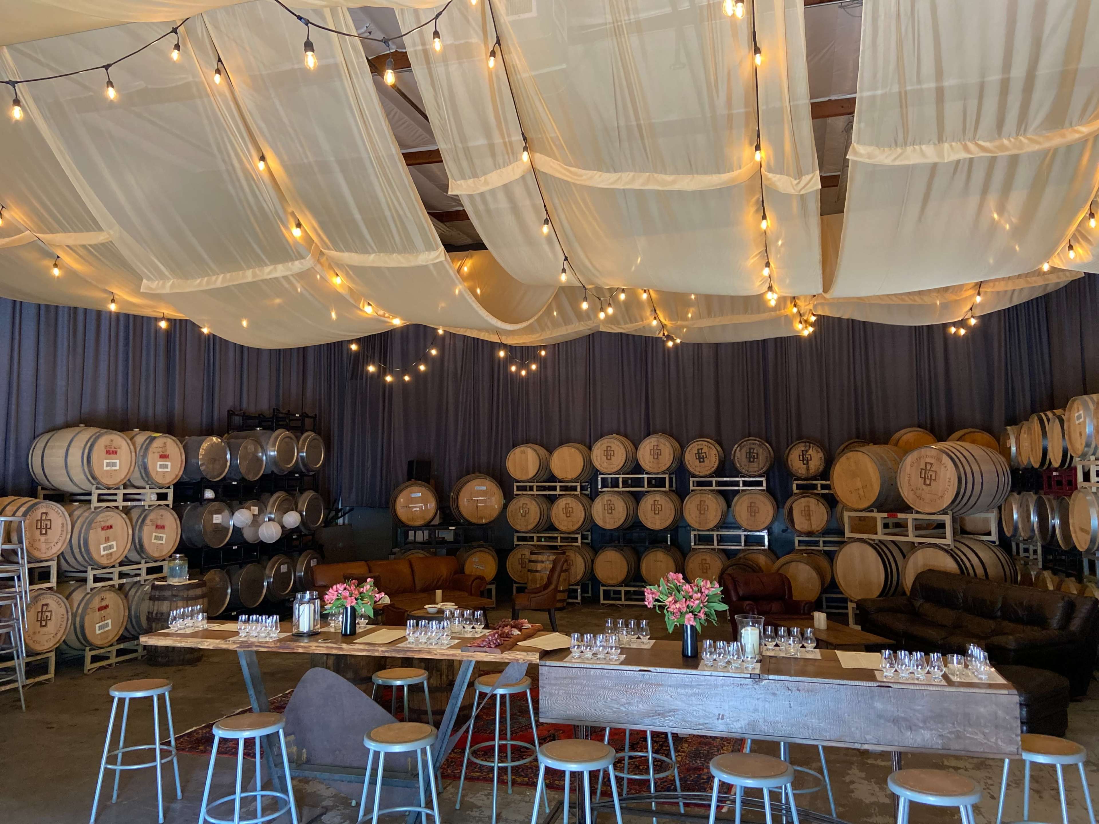 A winery tasting room with wooden barrels lining the walls, a long table set with glassware, and string lights hanging from a draped ceiling.