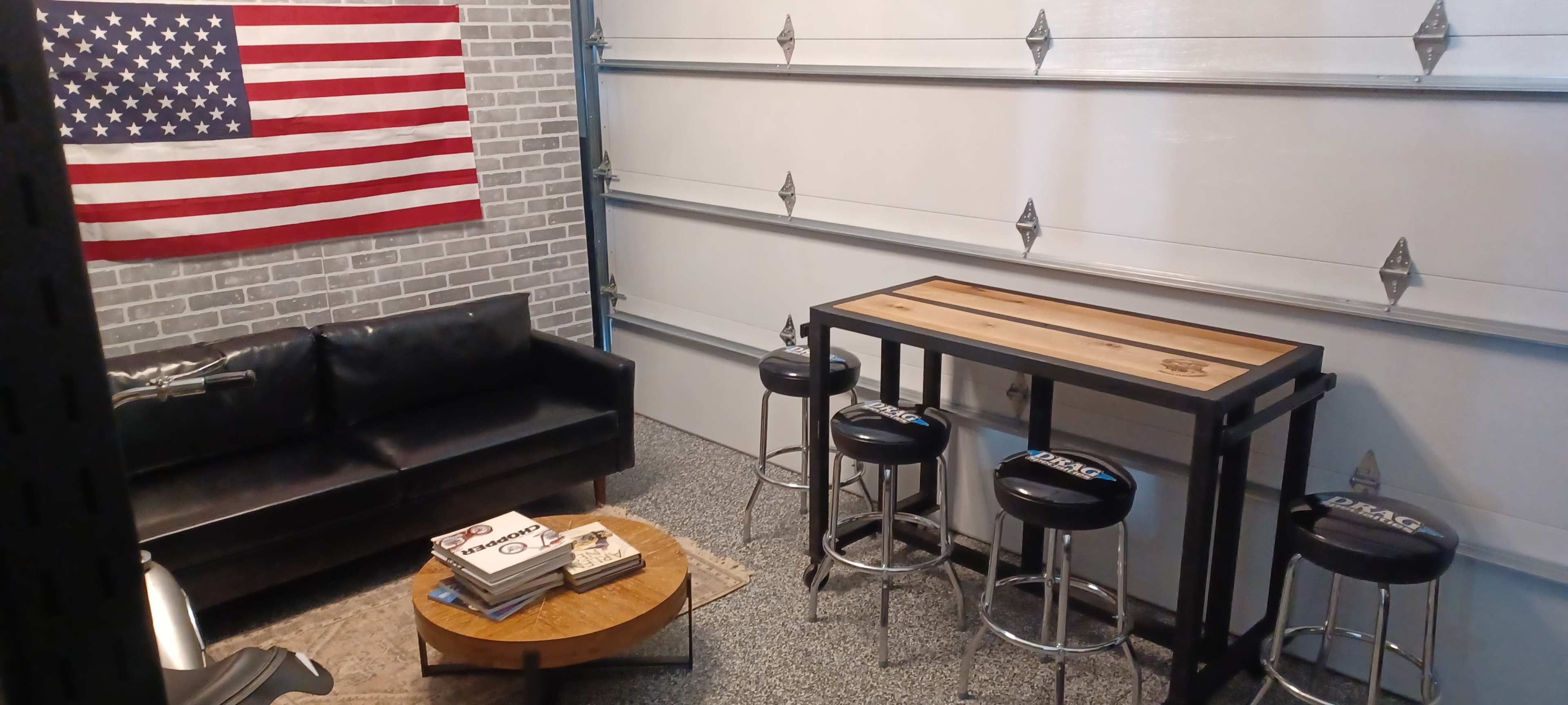 A garage space featuring a black couch, a wooden table with metal stools, a stack of magazines, and an American flag hanging on the wall.