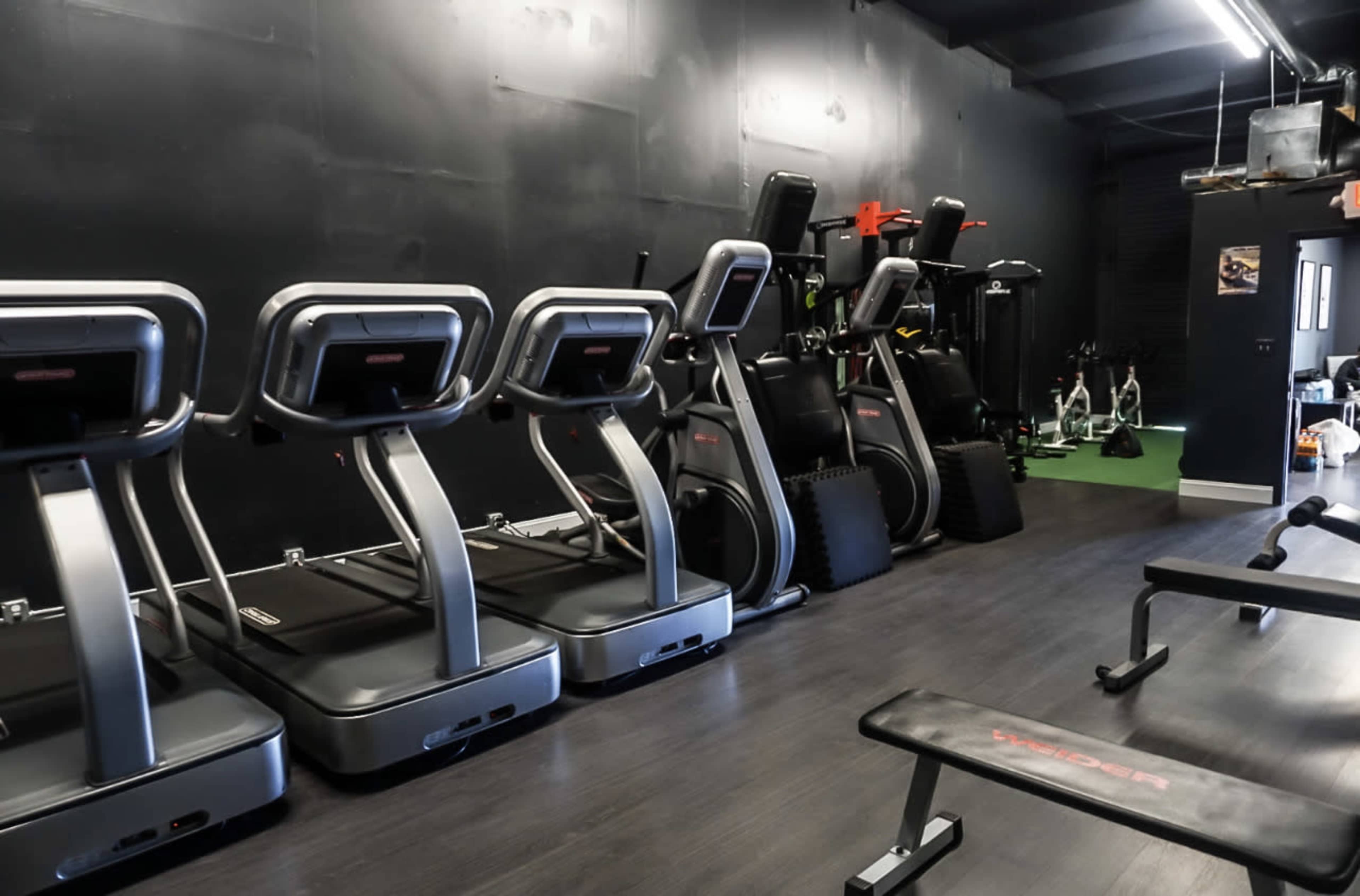 A fitness space featuring several treadmills lined up against a black wall, along with strength training equipment in the background.