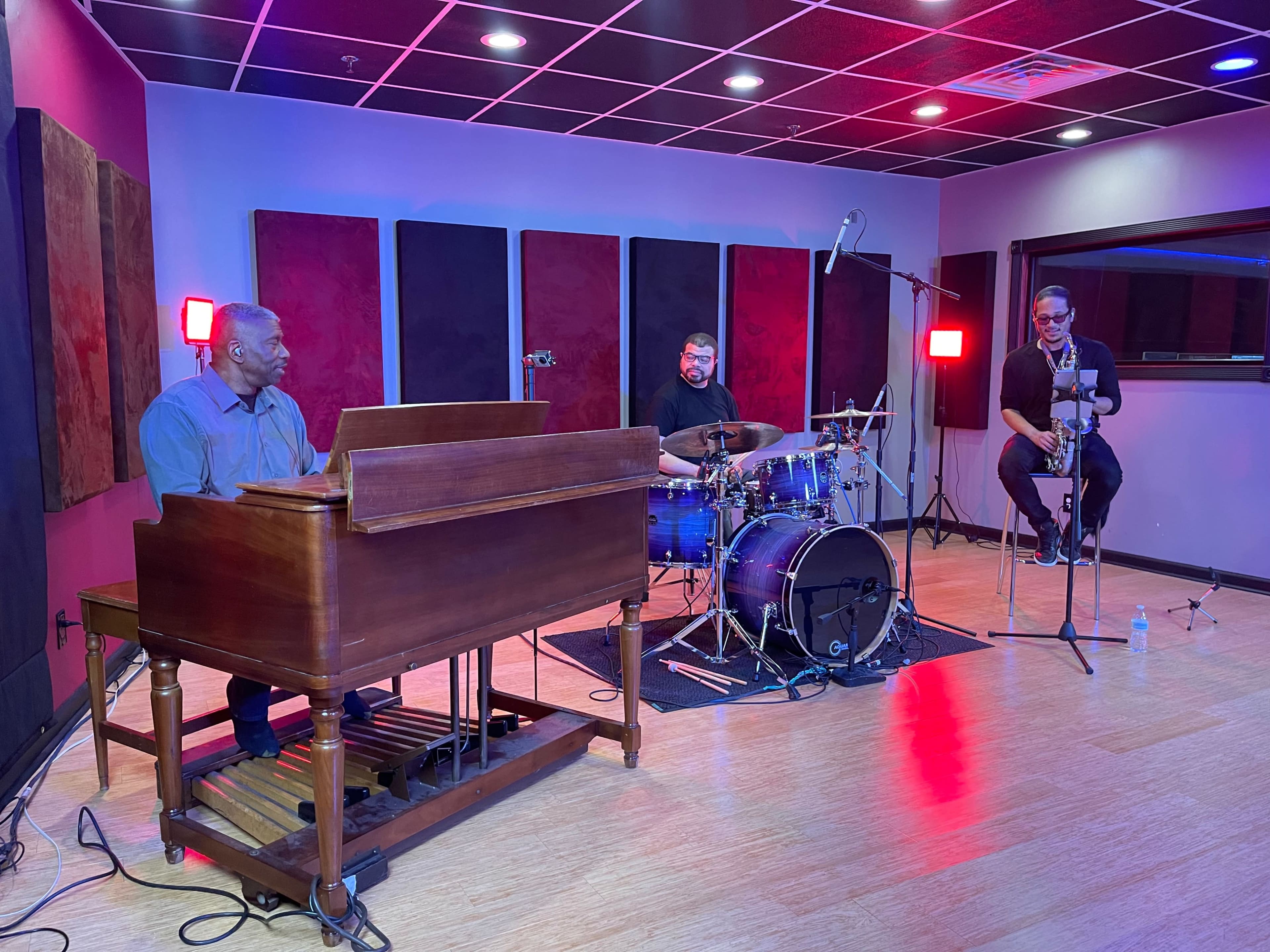 Three musicians perform in a recording studio, with a pianist on an organ, a drummer behind a drum set, and a saxophonist seated nearby.