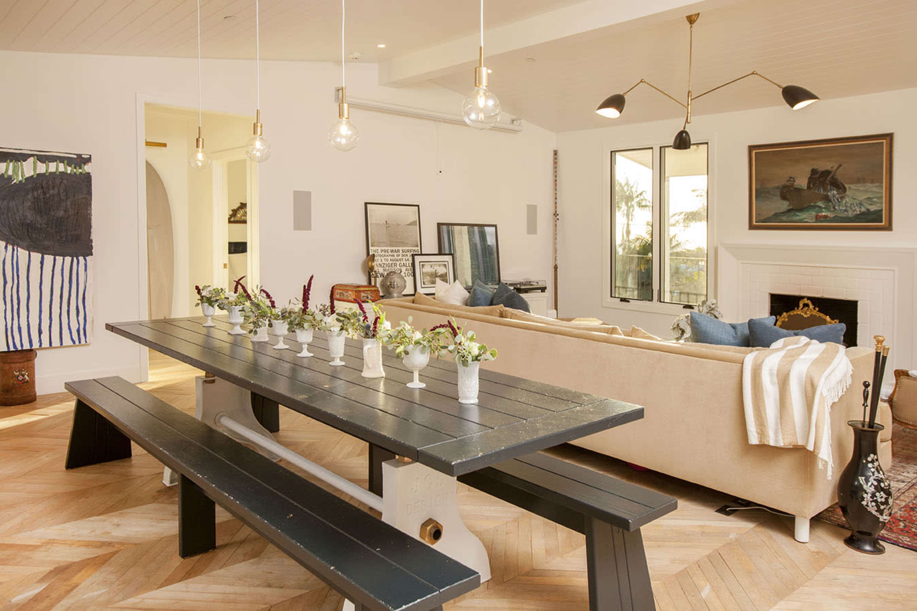 The living space features a long dark wooden table with small floral arrangements, surrounded by a beige couch and various artworks on the walls.