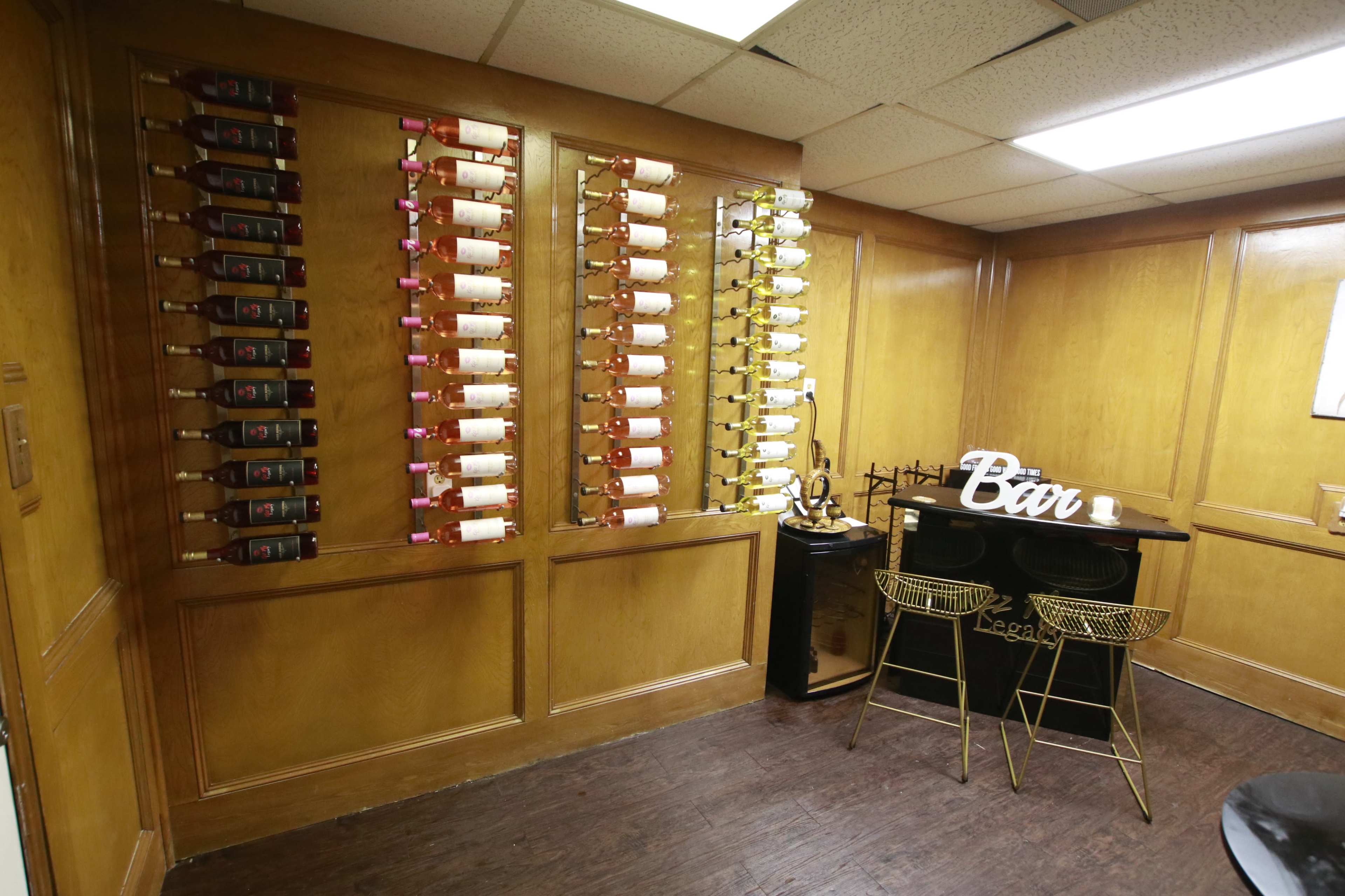 A room with wooden paneling featuring a wine rack on one wall filled with bottles, and a small bar area with gold-colored chairs and a decorative "Bar" sign.