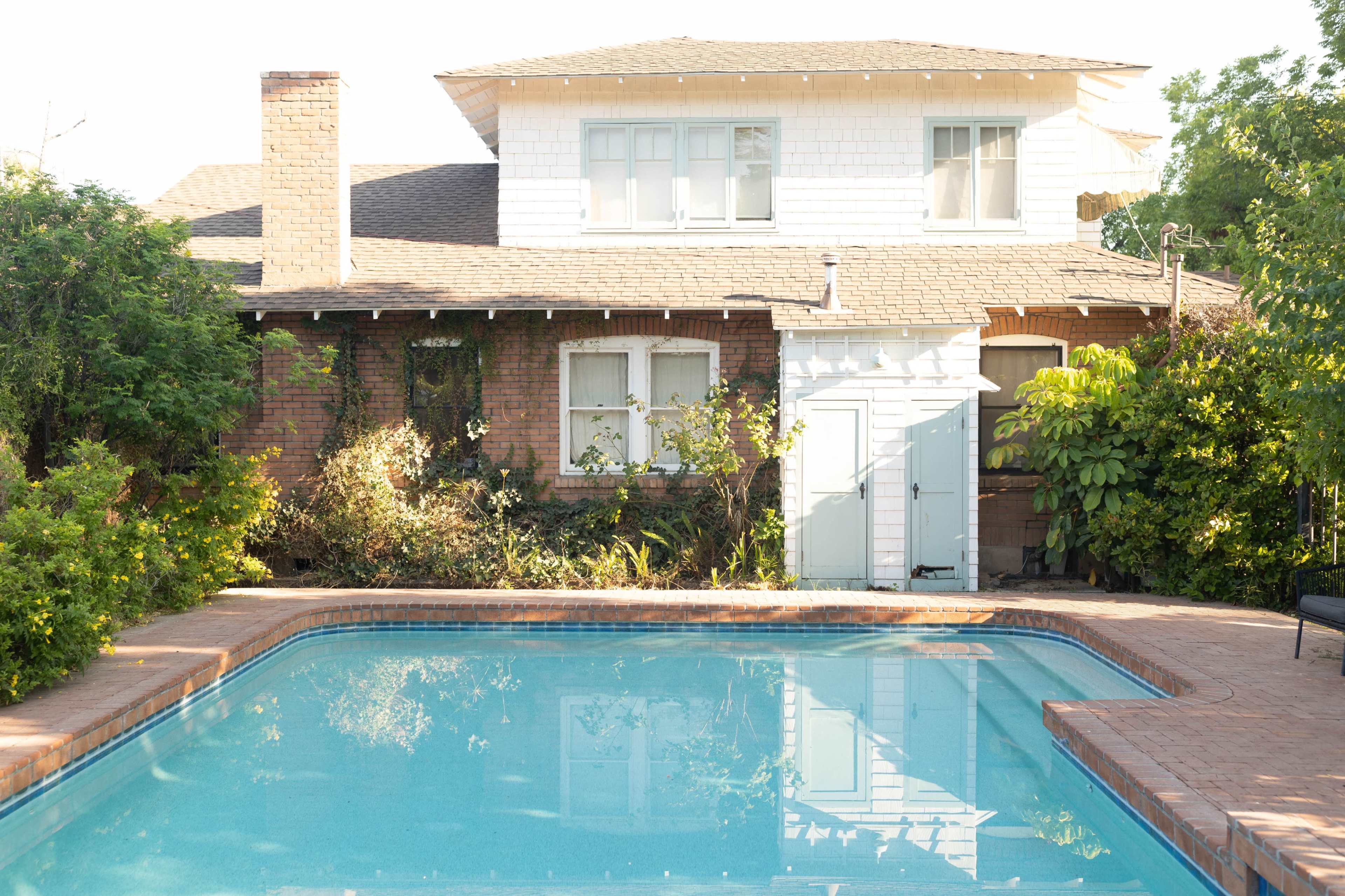 A clear swimming pool in front of a two-story house with a mix of brick and white siding, surrounded by lush greenery.