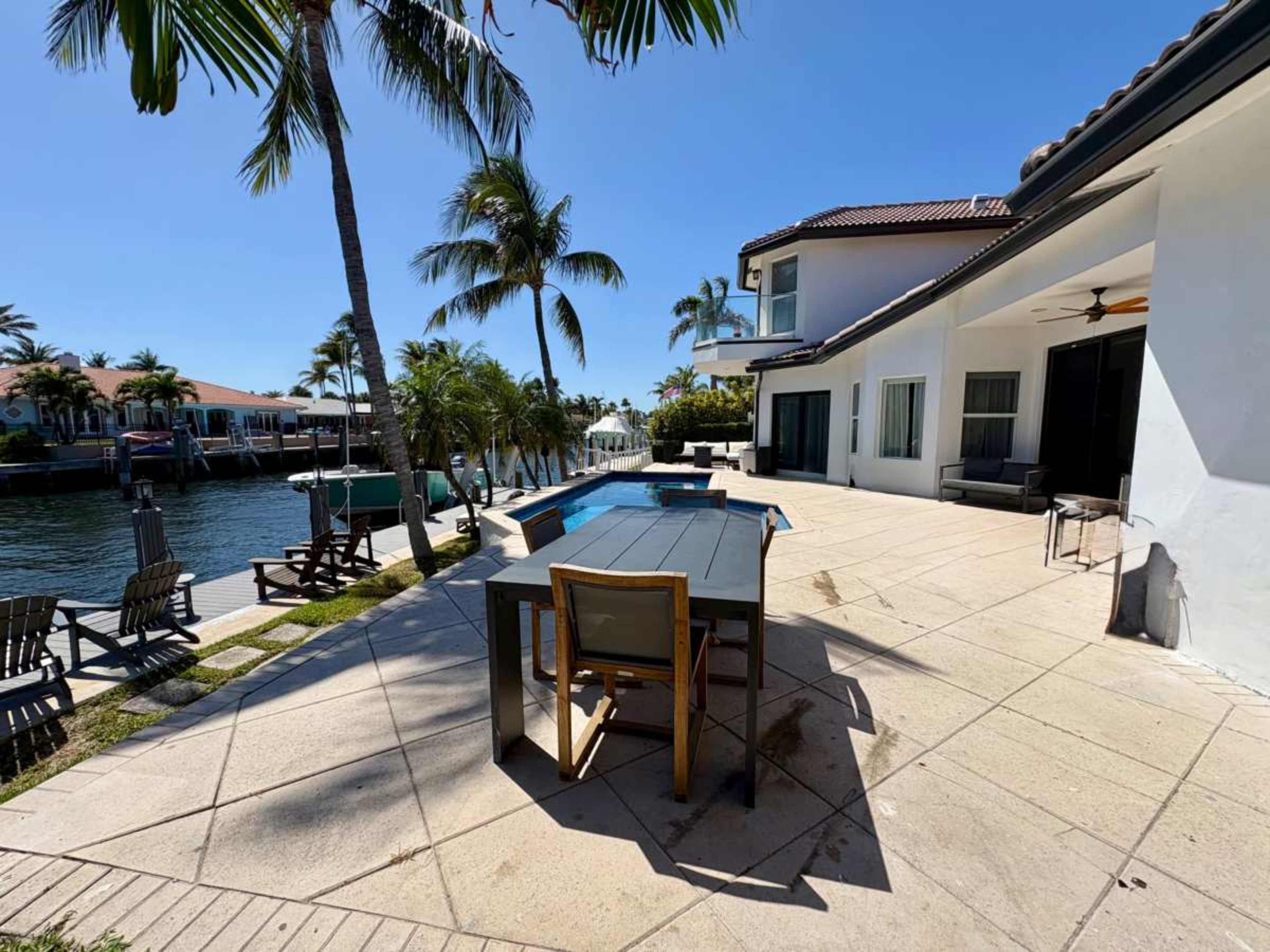 The image shows a waterfront patio featuring a pool and outdoor dining area, surrounded by palm trees and houses along a canal.