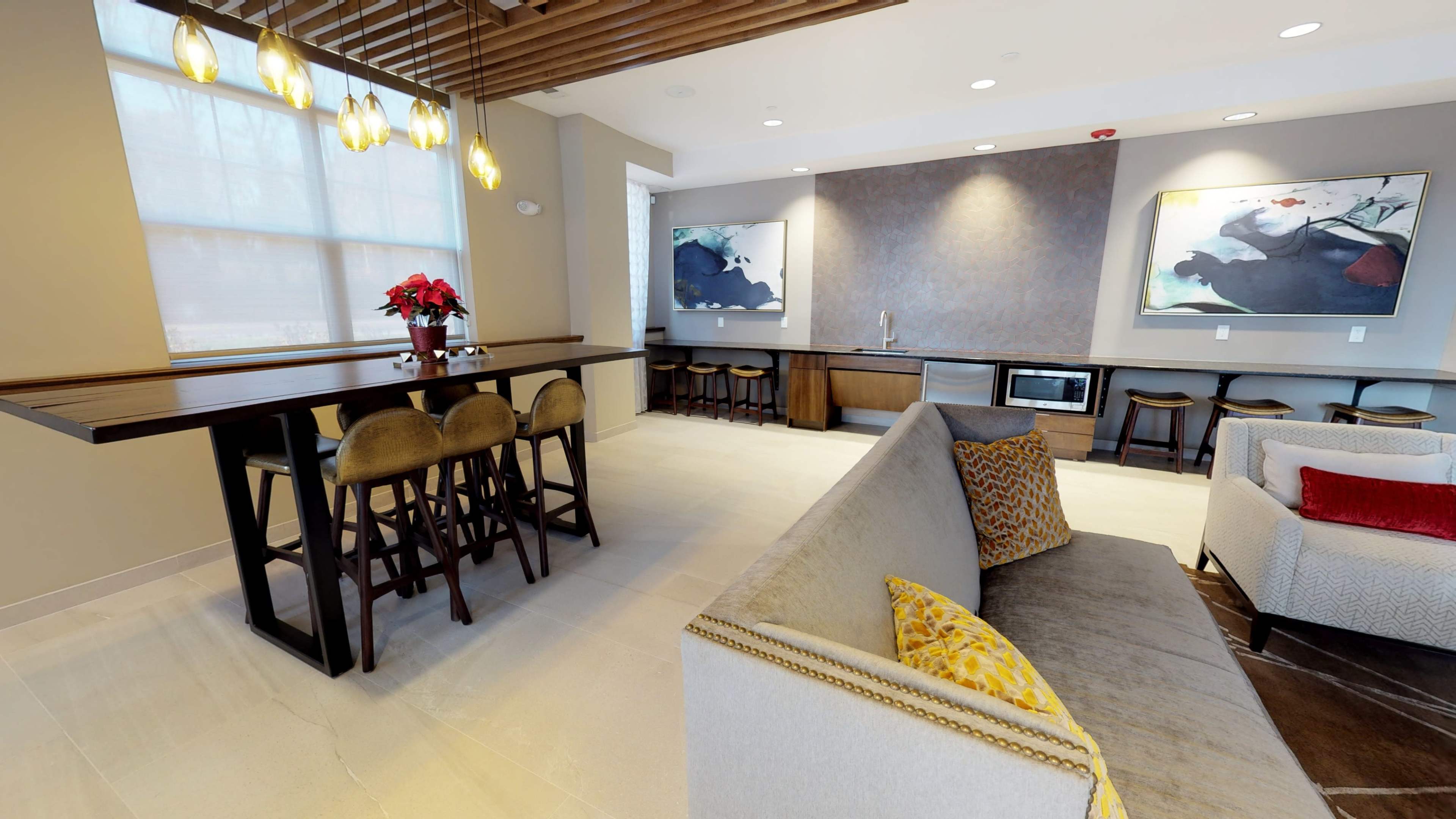 The image shows a modern lounge area featuring a long wooden table with bar stools, a kitchenette, and a comfortable seating arrangement.