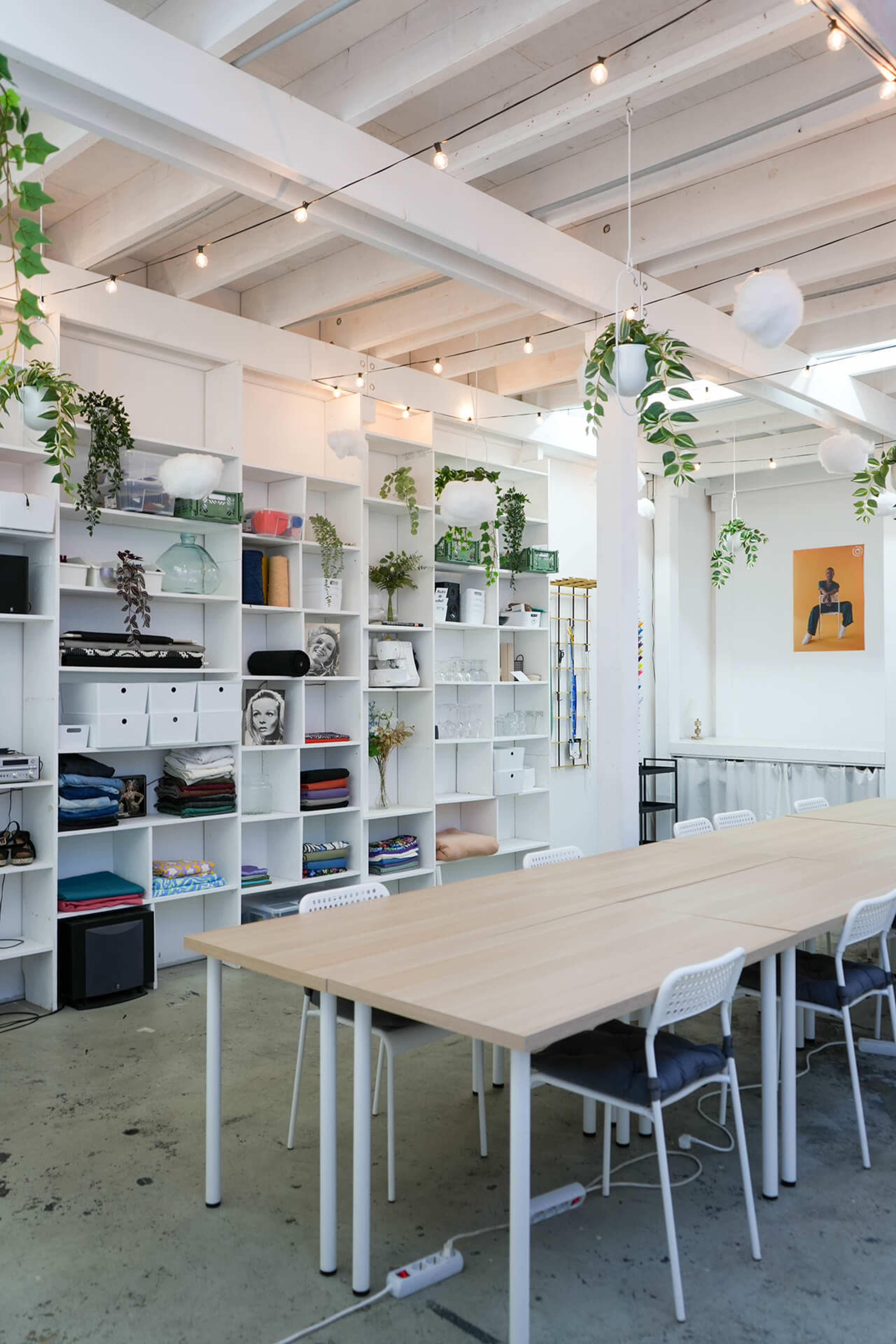 A modern workspace features a large wooden table surrounded by white chairs, set against a backdrop of organized shelves filled with various items and greenery.