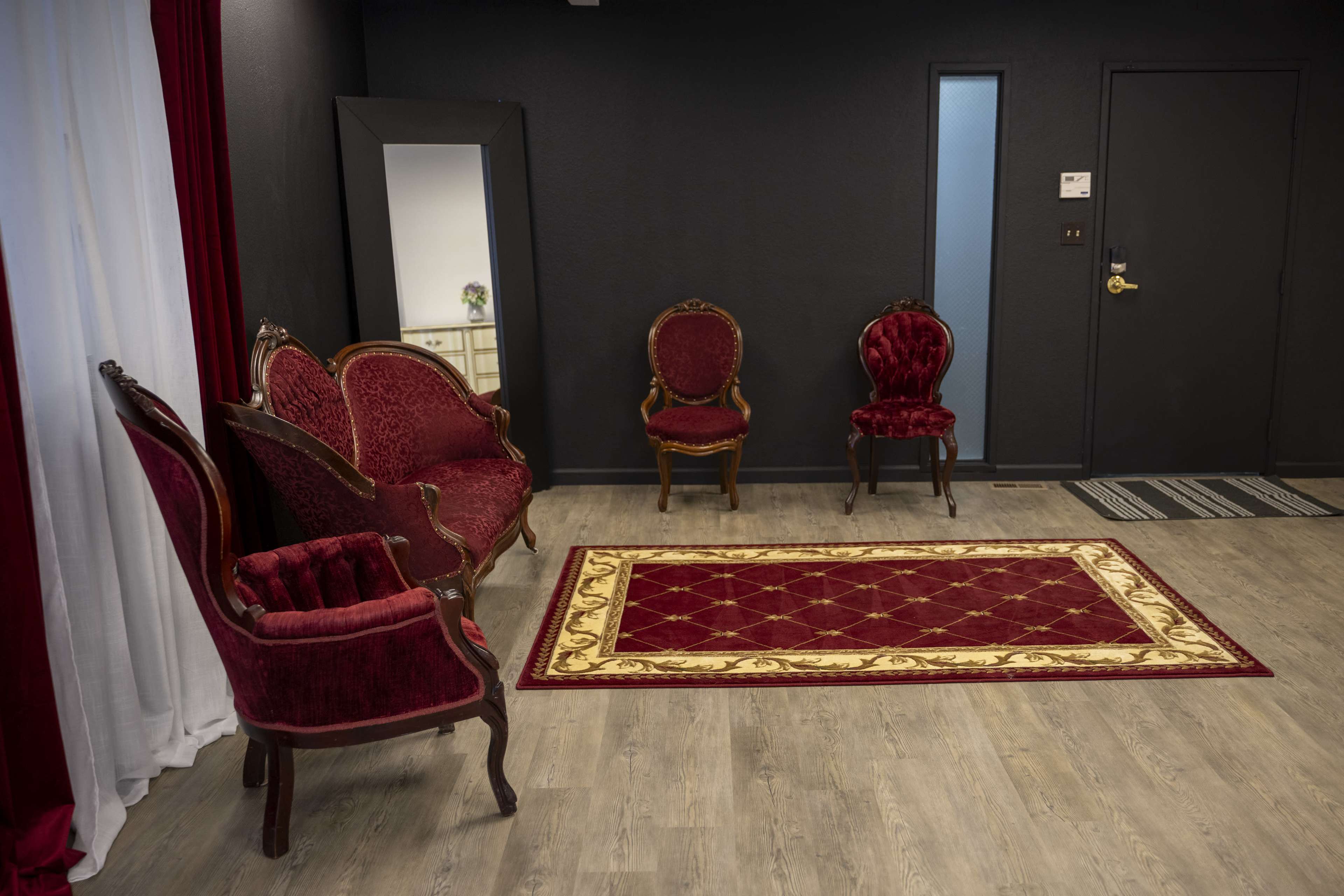 A room with a red carpet, two ornate red upholstered chairs, and a mirror, with a door visible in the background.
