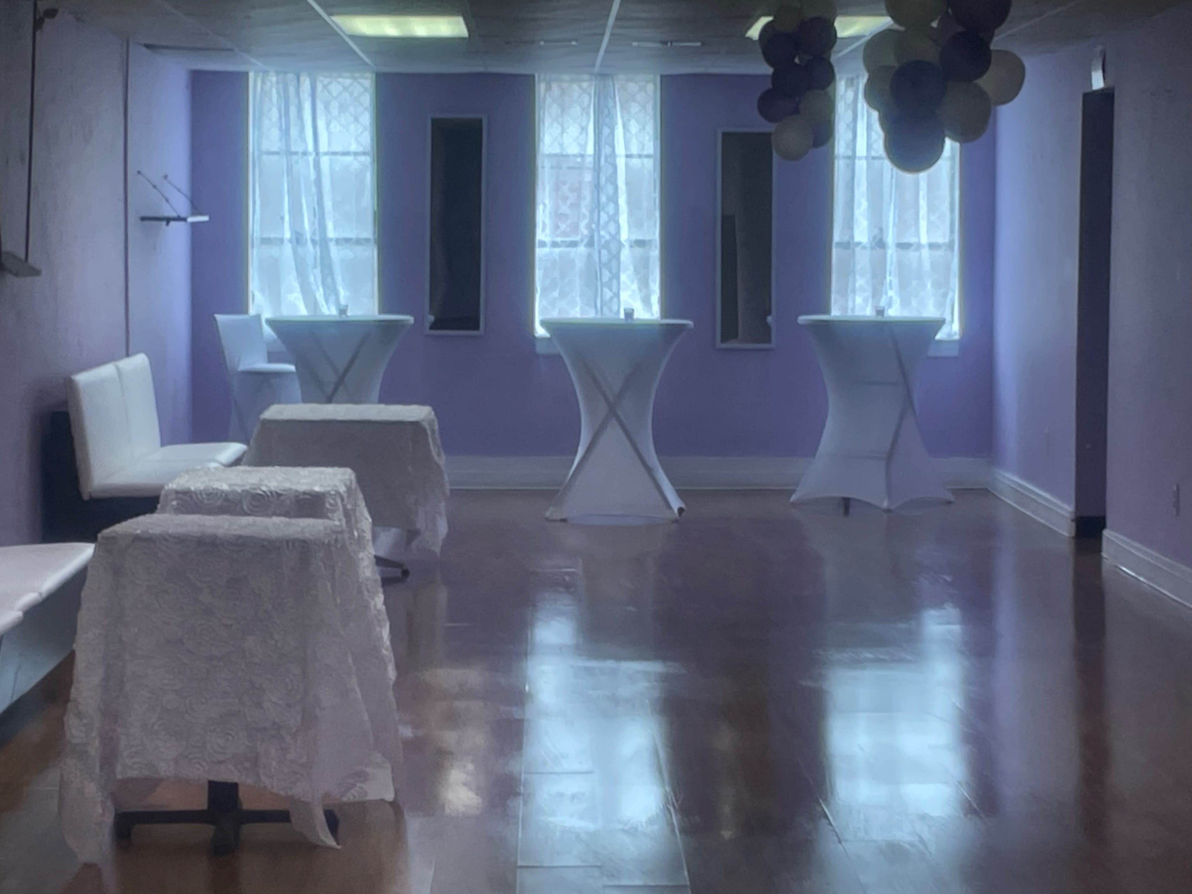 A spacious room with purple walls, equipped with several white tables draped in tablecloths and illuminated by natural light from large windows.