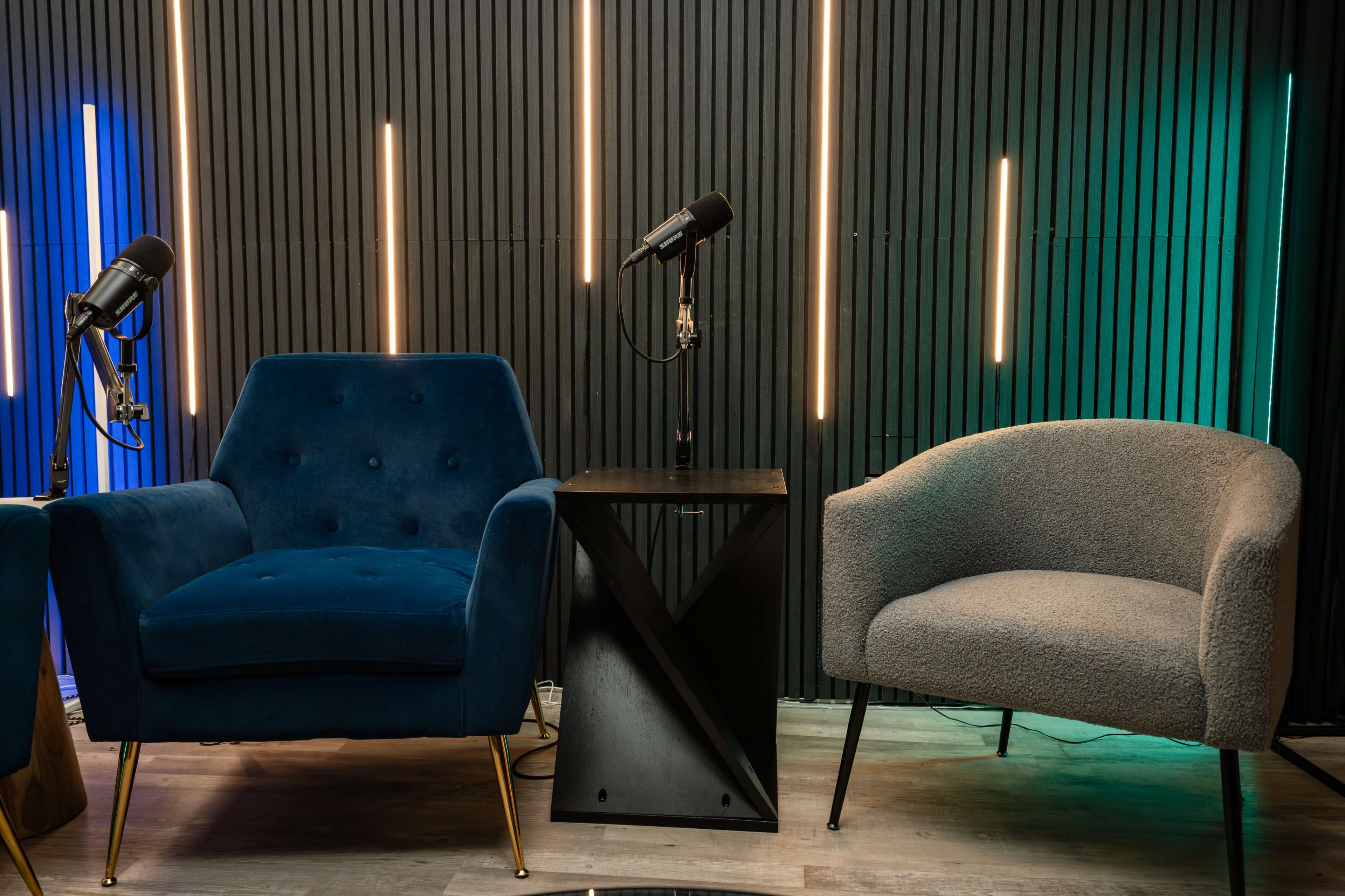 The image shows two chairs, one blue and one gray, situated next to a small black table, with microphones positioned nearby against a backdrop of vertical light strips.