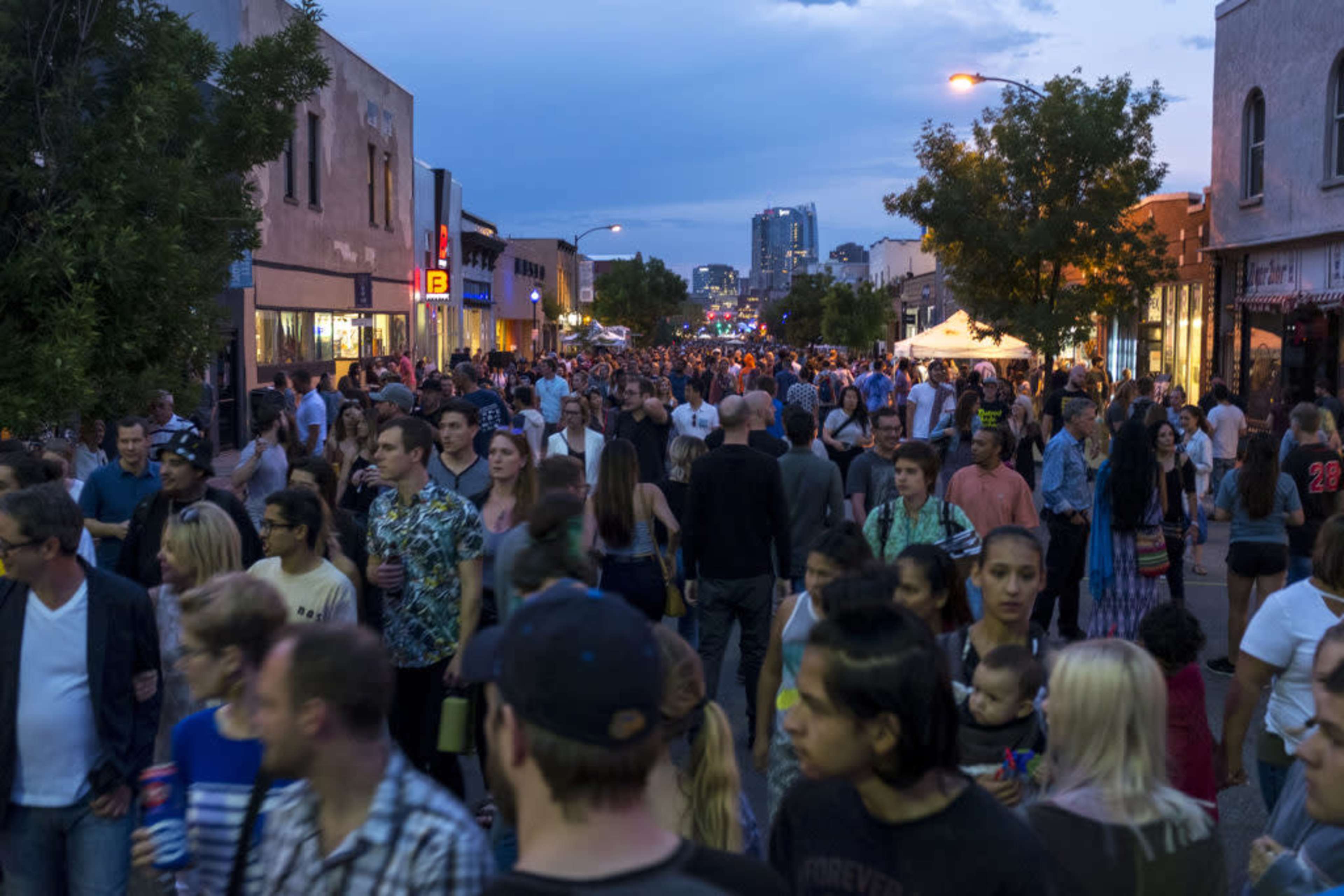 A large crowd of people fills a street with shops and buildings, under a dusk sky.