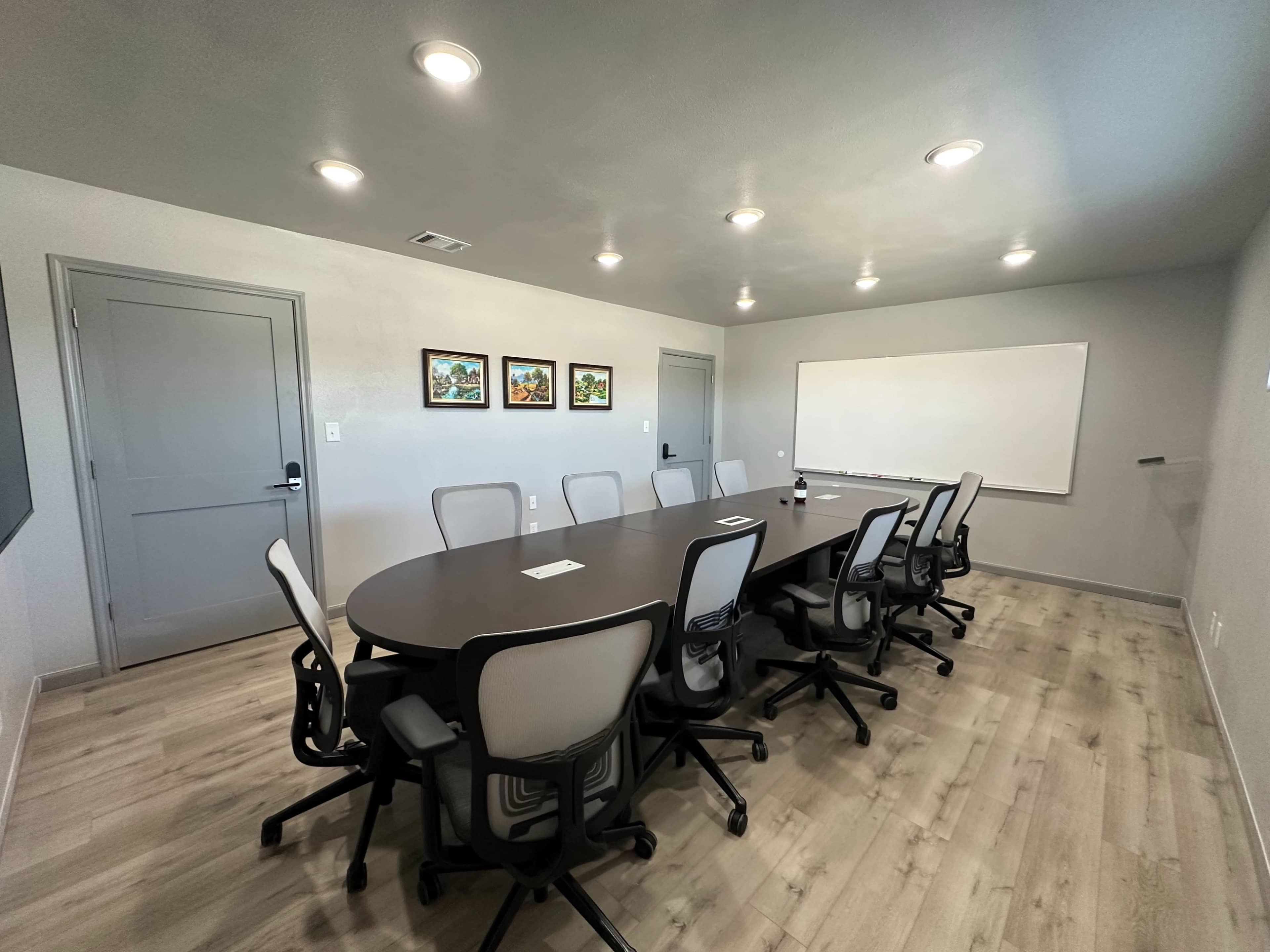 A modern conference room features a large oval table surrounded by ergonomic chairs, with a whiteboard and framed artwork on the walls.