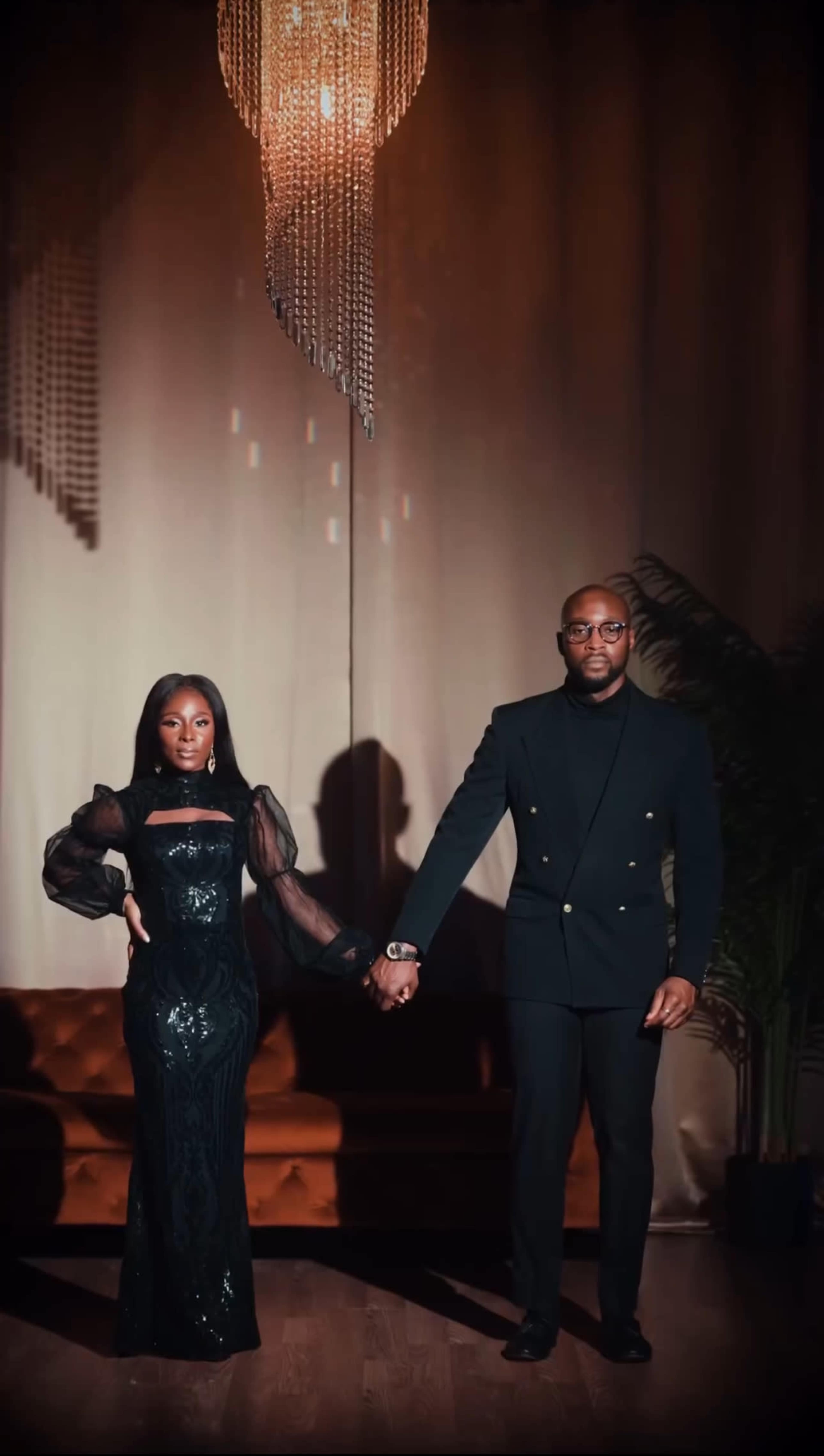A man and woman stand hand in hand on a stage with a chandelier above and a subtle backdrop.