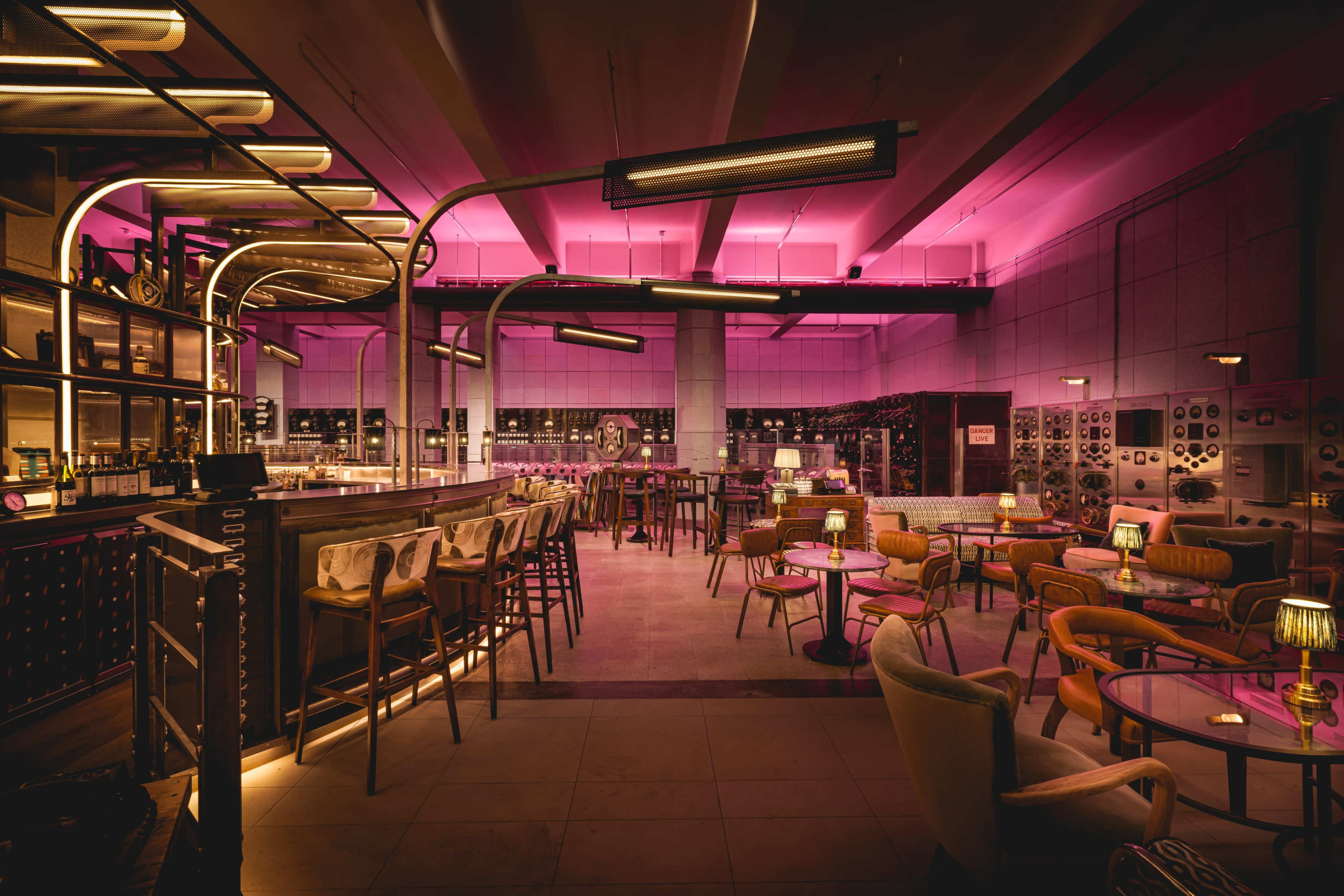 A stylish bar interior with neon pink lighting, featuring various seating arrangements and a long counter.