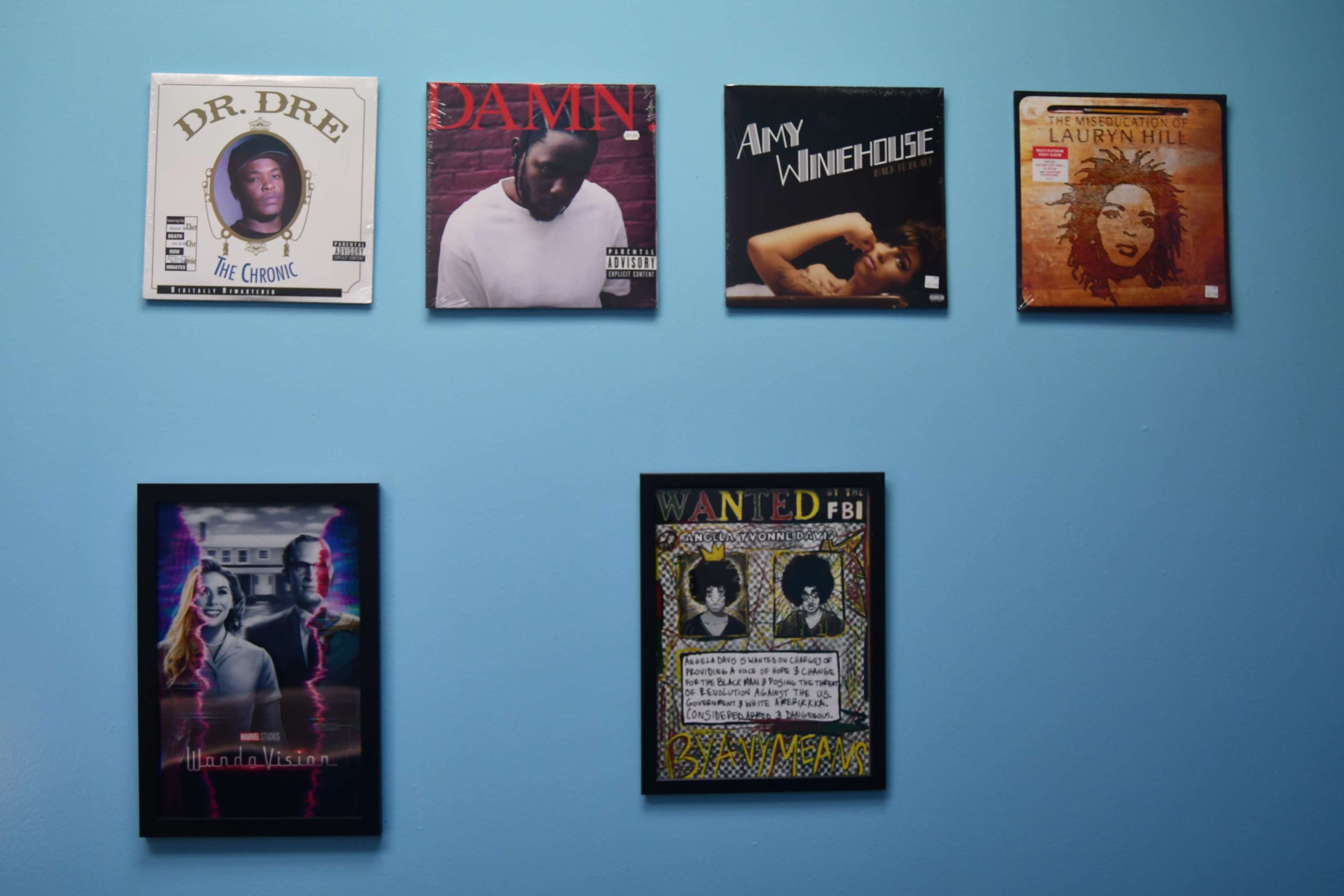 A blue wall featuring framed album covers of various artists, including Dr. Dre, Kendrick Lamar, Amy Winehouse, and Lauryn Hill, along with a framed poster labeled "WANTED" with a stylized design.