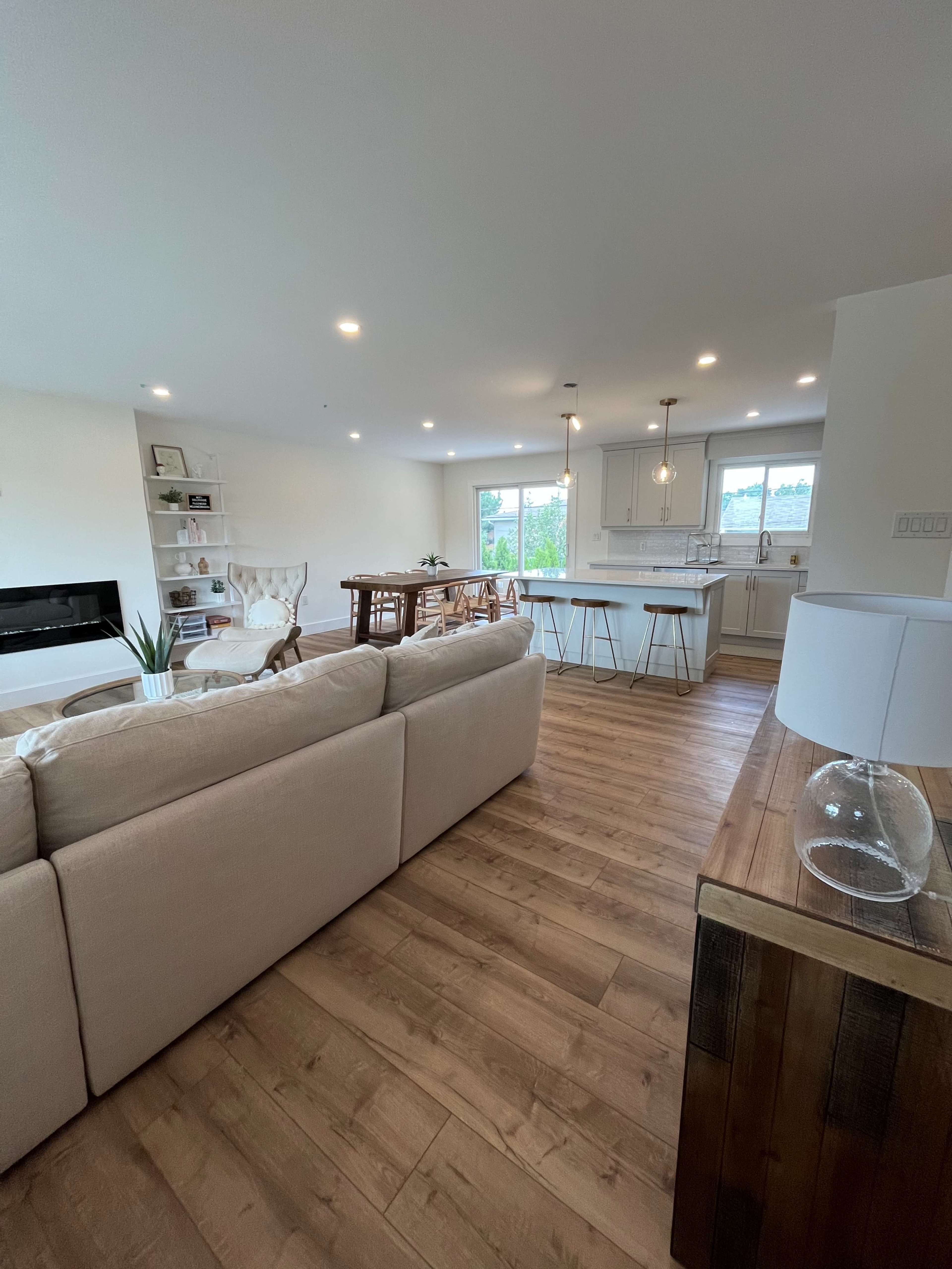 A modern living space with a beige sofa, a dining area with a wooden table, and a kitchen in the background, featuring white cabinetry and pendant lighting.