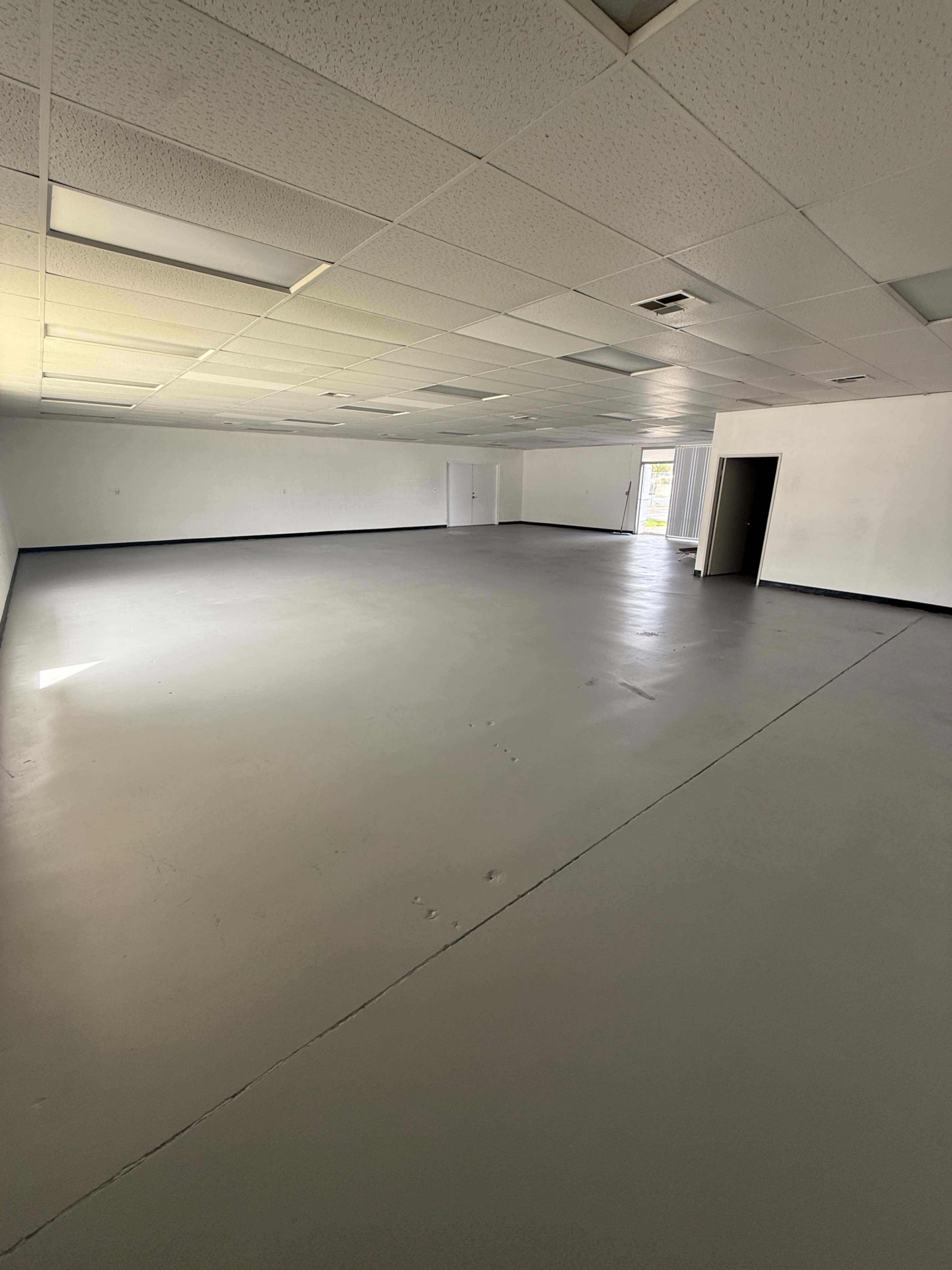 The image shows a large, empty room with concrete flooring, overhead fluorescent lights, and a single door on one side.