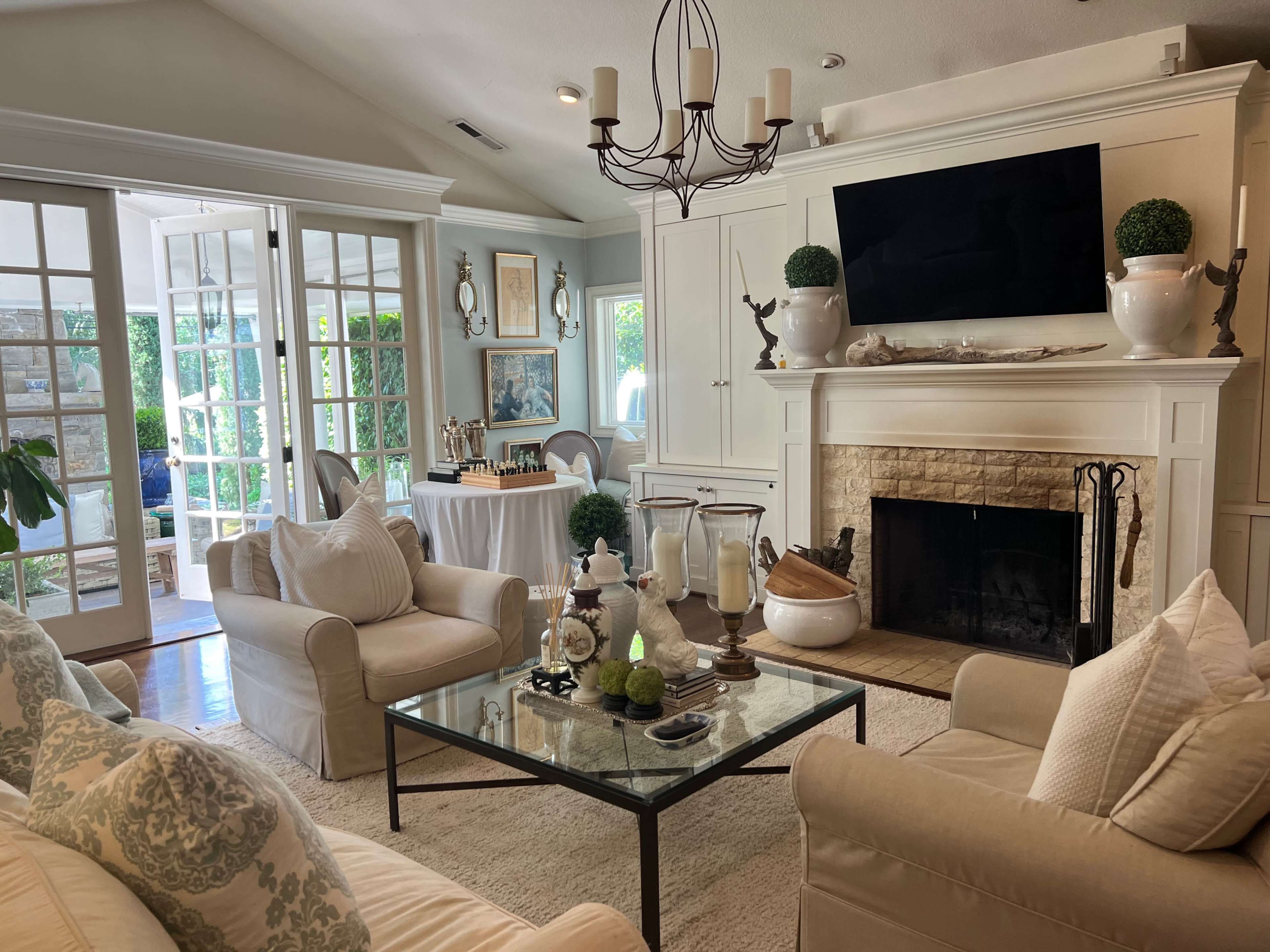 A light-filled living room with a cream-colored sofa, a glass coffee table, and a stone fireplace adorned with decorative elements, leading to a sunlit sunroom through French doors.